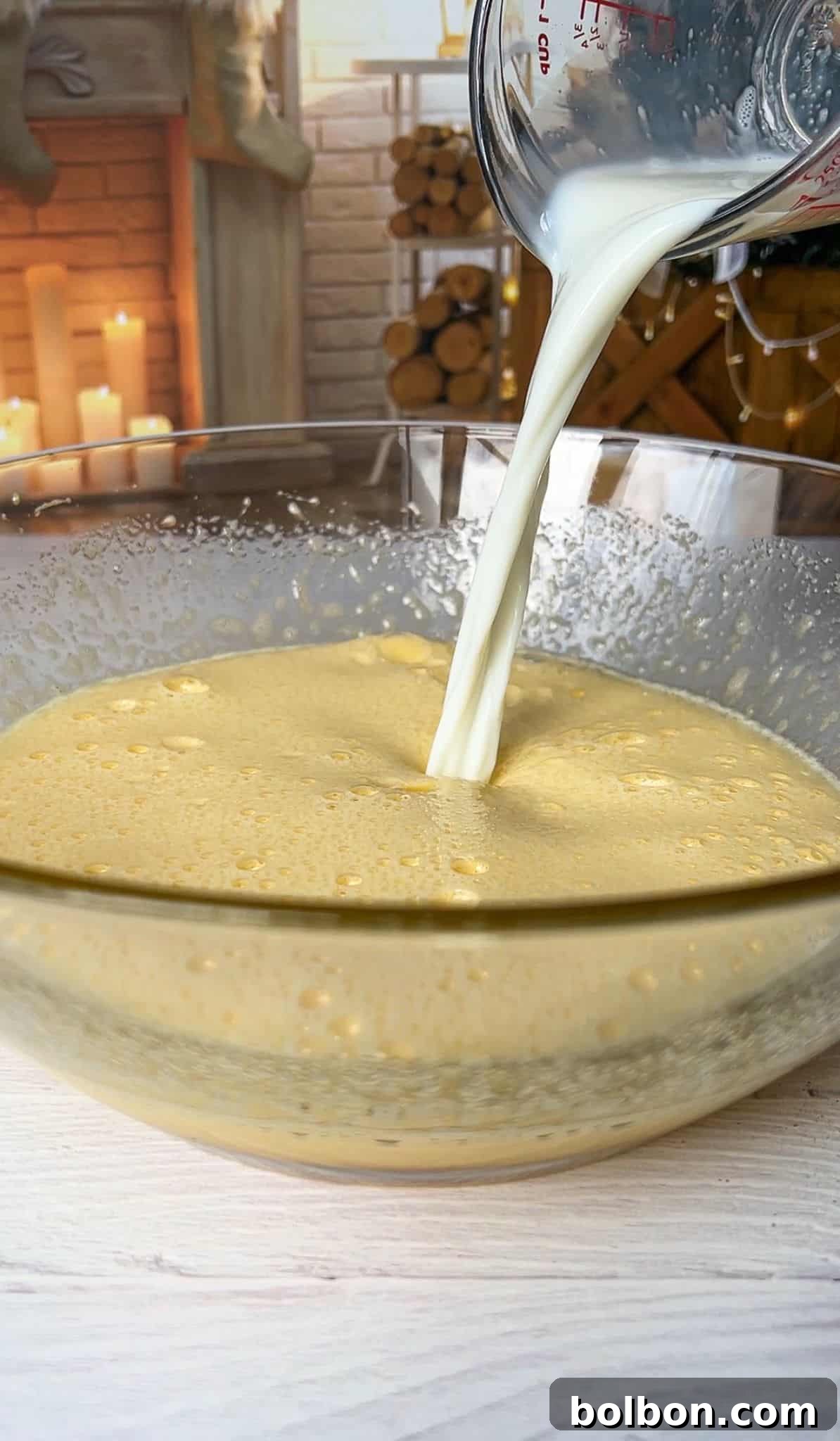 The Elusive Apple Cake 8 Milk being poured into a glass bowl filled with a frothy yellow mixture. In the background, a rustic setting features stacked firewood and lit candles beside a brick fireplace.