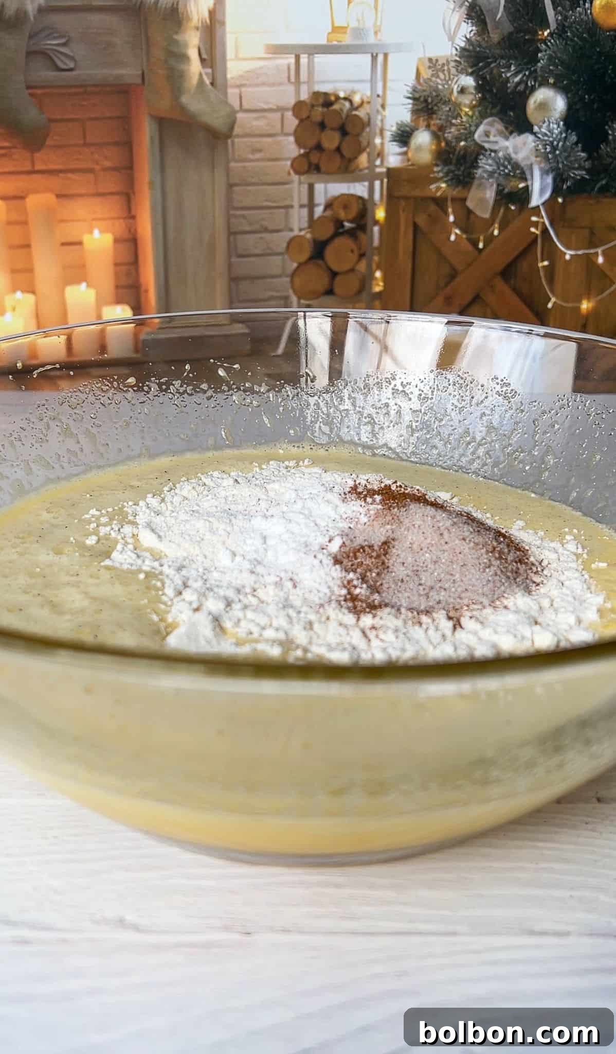 The Elusive Apple Cake 9 A glass bowl filled with cake batter and topped with flour and cocoa sits on a white kitchen counter. In the background, a decorated Christmas tree and a lit fireplace are visible, creating a cozy holiday atmosphere.