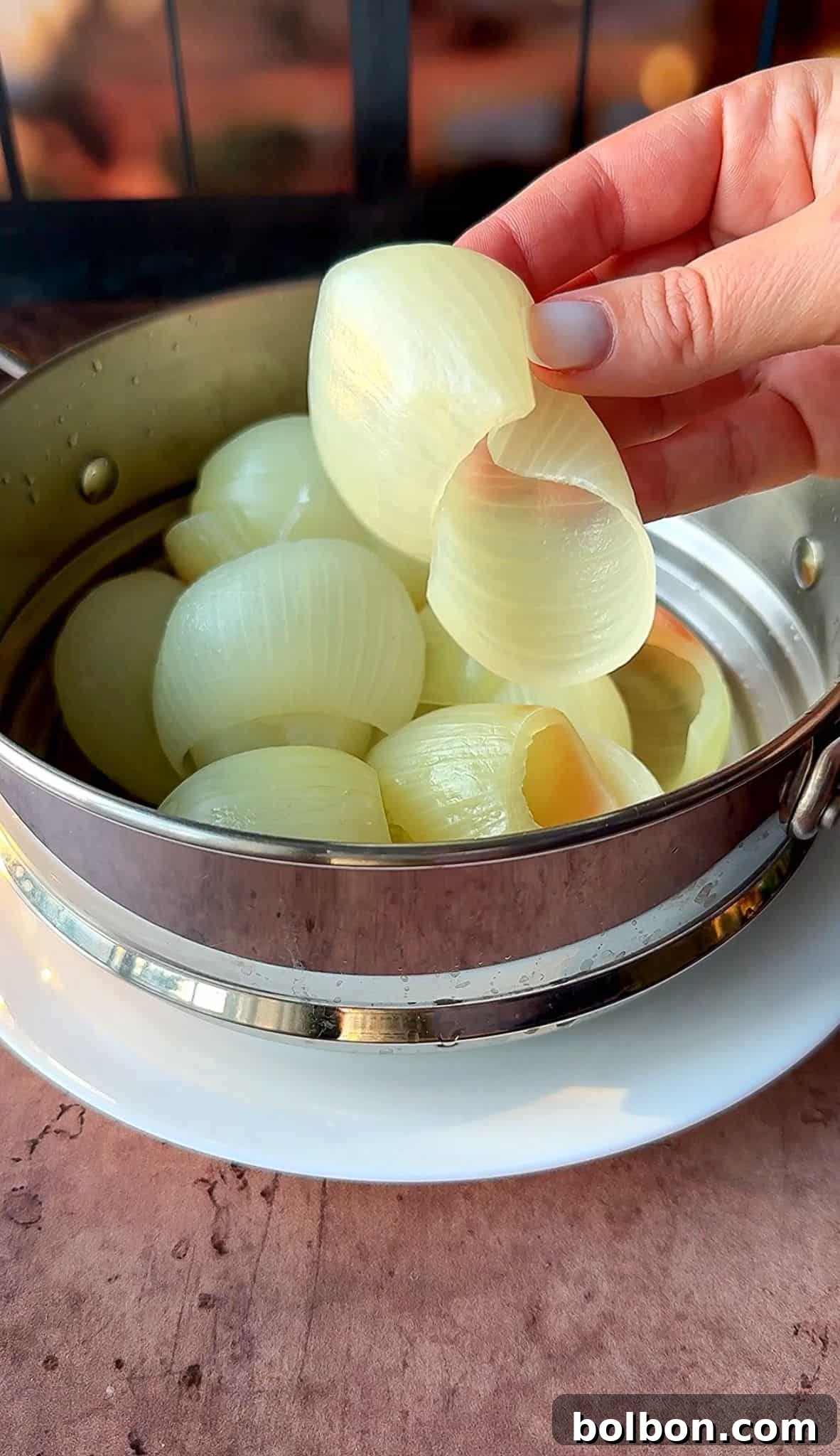 Golden Stuffed Onion Delights 11 A hand peeling layers from a partially transparent onion in a stainless steel pot. The pot rests on a white plate. The background is blurred, focusing attention on the onion layers and hand.