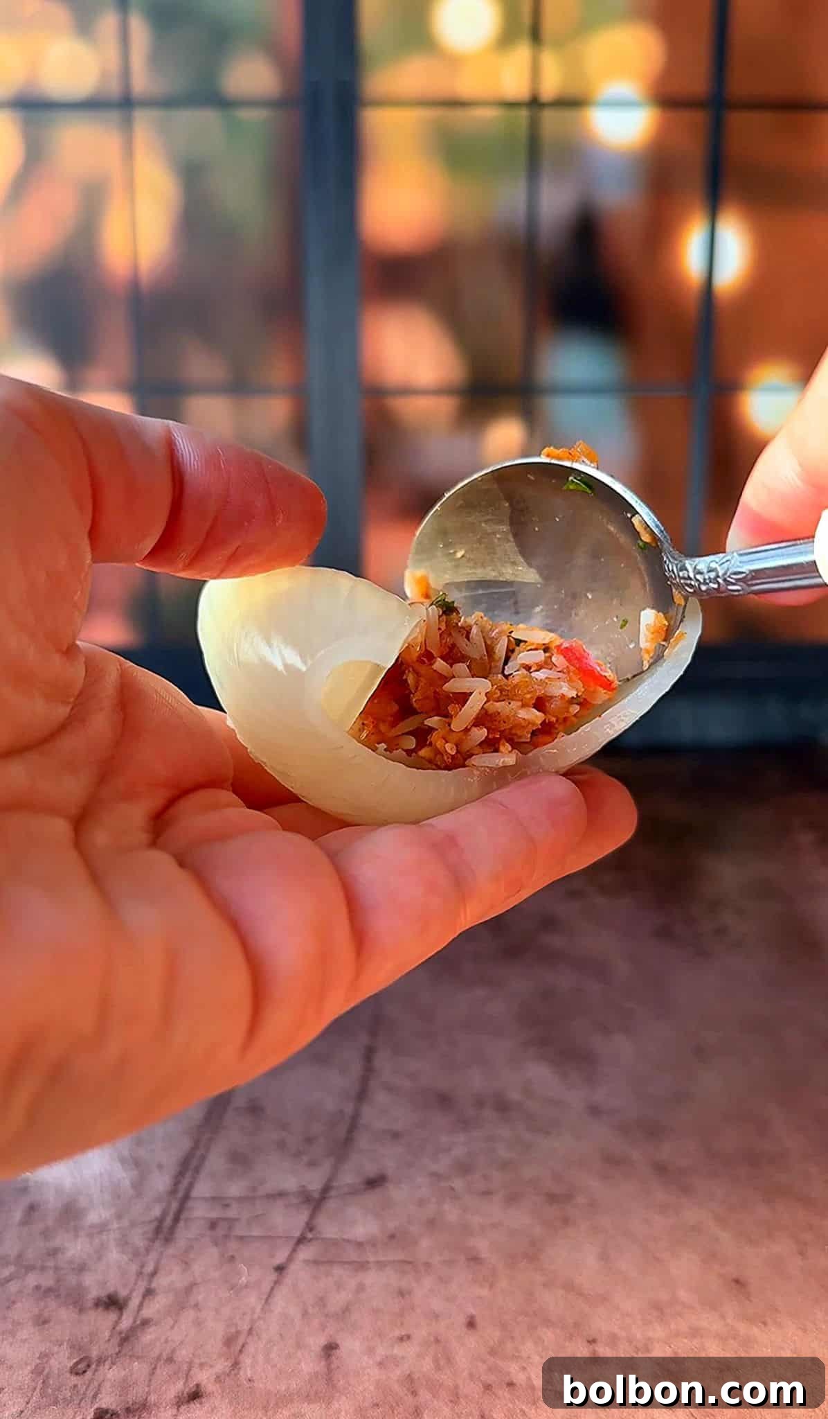 Golden Stuffed Onion Delights 12 A hand holding a piece of raw onion being filled with a mixture of rice and vegetables using a spoon. The background is softly focused, with warm lighting.