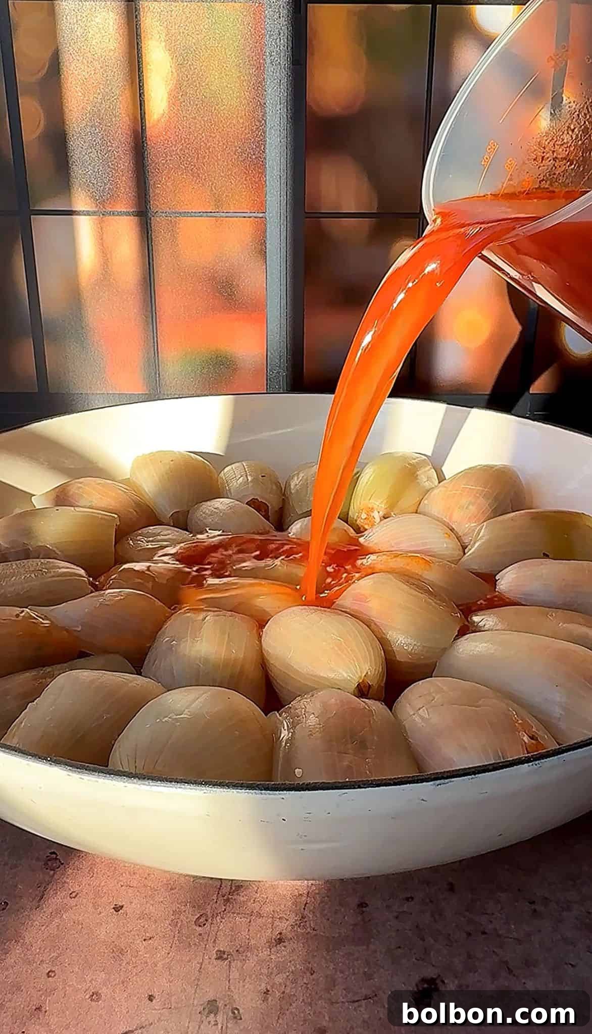 Golden Stuffed Onion Delights 14 A bowl filled with halved onions is being drenched with a vibrant red sauce poured from a container, against a backdrop of warm, blurred lighting and window shadows.