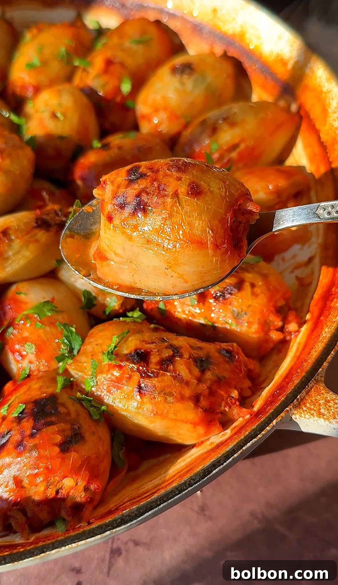 Golden Stuffed Onion Delights 18 A spoon holding a stuffed onion with a golden-brown surface, garnished with herbs. The dish is filled with similar stuffed onions, cooked and slightly charred, served in a rustic pan.