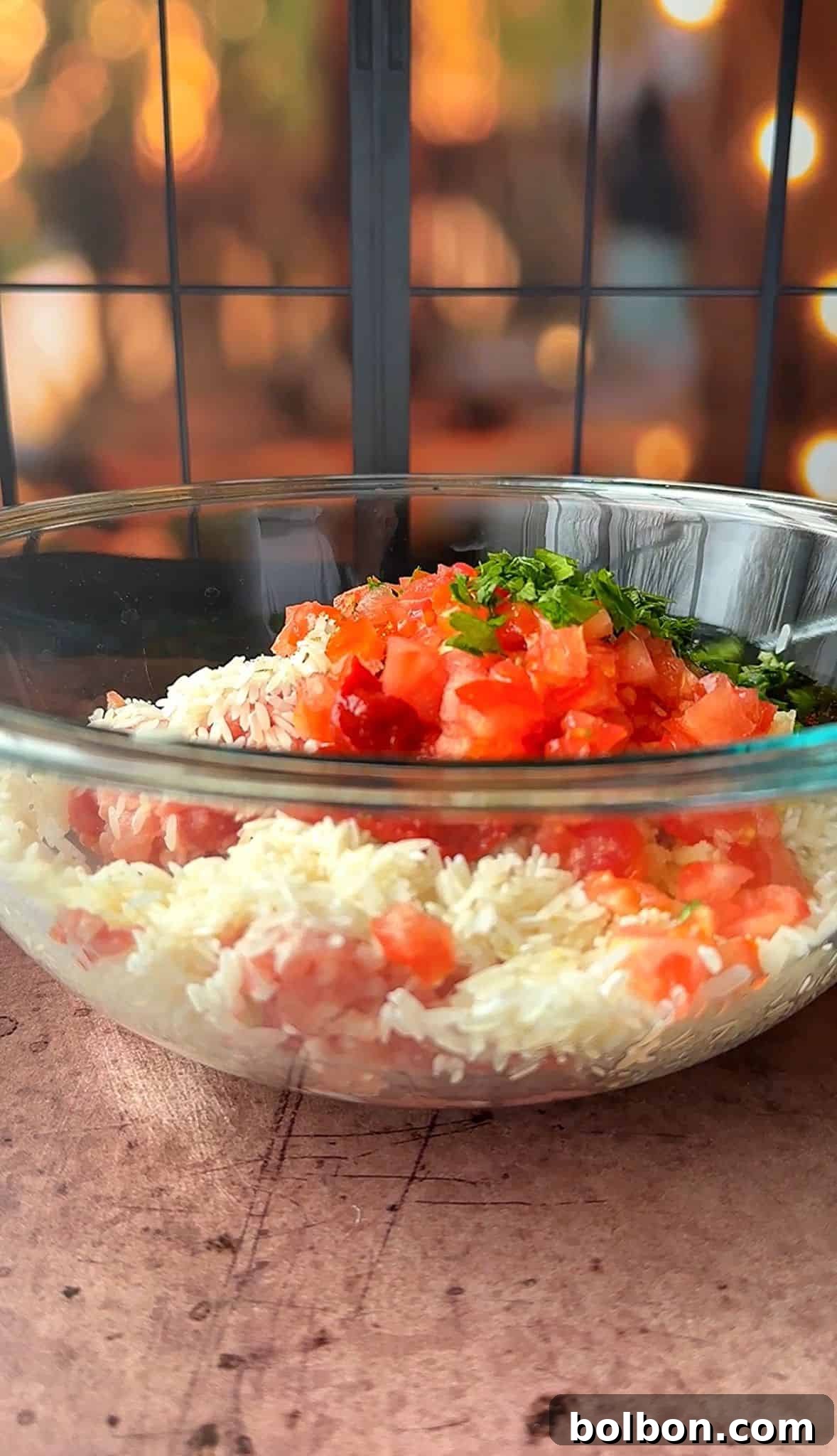 Golden Stuffed Onion Delights 4 A glass bowl filled with rice topped with diced tomatoes and chopped herbs. The background features a softly lit window with warm, blurred lights.
