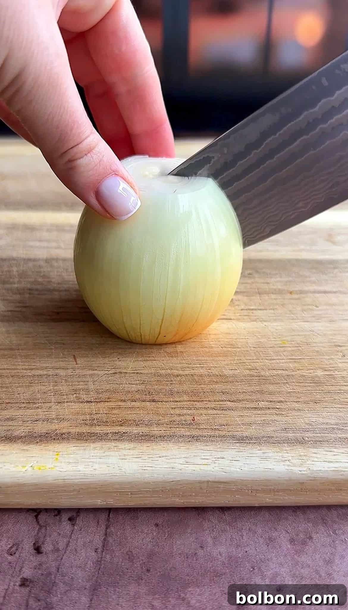 Golden Stuffed Onion Delights 7 Close-up of a person’s hand holding an onion on a wooden cutting board, while a knife is poised about to slice the onion. The background is softly blurred.