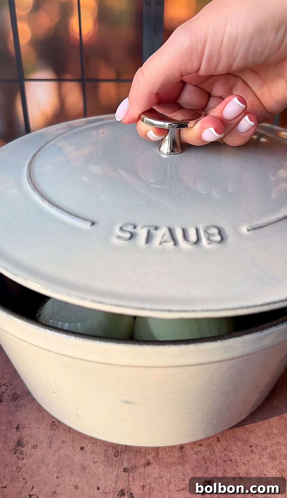 Golden Stuffed Onion Delights 9 A hand with manicured nails lifts the lid of a Staub Dutch oven, revealing onions inside. The scene is set on a wooden surface with a blurred backdrop, suggesting an outdoor or patio setting.