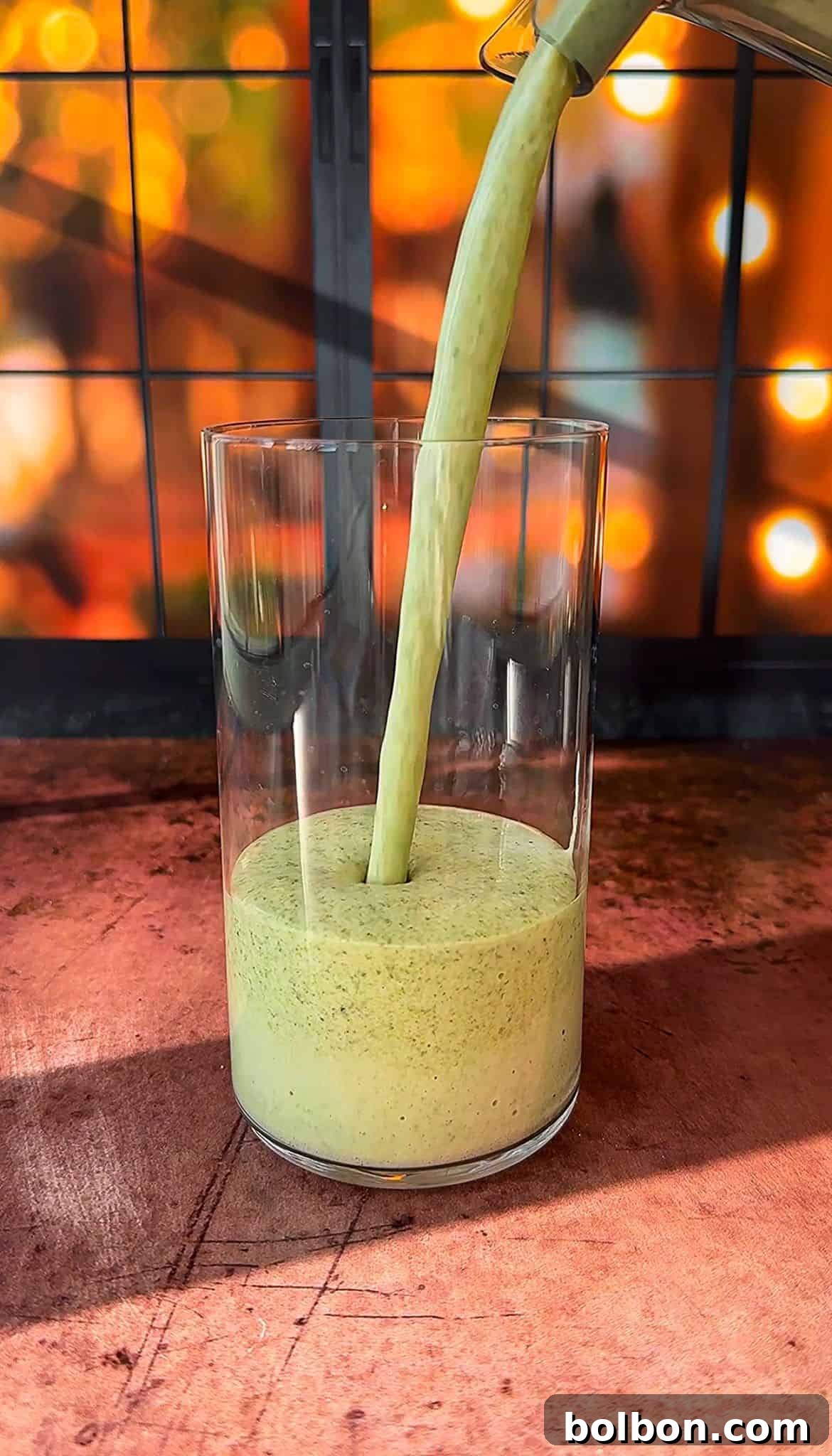 A thick, green smoothie is being poured into a clear glass. The background features a warm, out-of-focus display of glowing lights, creating a cozy ambiance. The scene is set on a wooden surface.