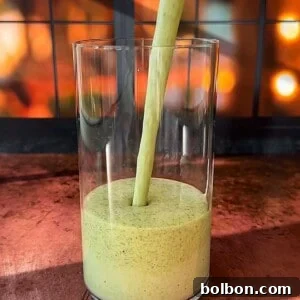 A thick, green smoothie is being poured into a clear glass. The background features a warm, out-of-focus display of glowing lights, creating a cozy ambiance. The scene is set on a wooden surface.
