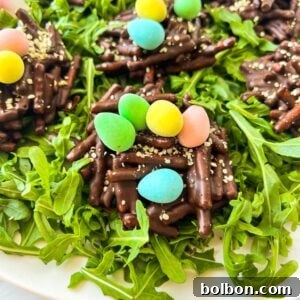 Chocolate nests topped with colorful candy eggs sit on a bed of fresh arugula. The nests are made of chocolate-coated sticks and sprinkled with sesame seeds. The eggs are pastel shades of blue, green, yellow, and pink.