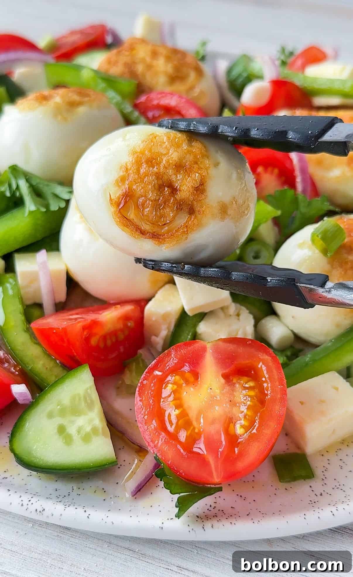 Crispy Golden Eggs 3 A close-up of a fresh, vibrant salad showcasing perfectly sliced cucumber, juicy cherry tomatoes, thinly cut red onion, crisp green pepper, and creamy cubes of feta cheese. A pair of tongs holds a beautifully fried boiled egg with a rich golden-brown top, garnished with fresh herbs, ready to be added to the salad.