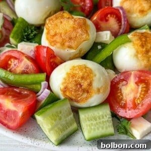 A colorful salad featuring grilled eggs, cherry tomatoes, cucumber slices, green pepper, red onion, and parsley on a white plate. The vegetables are fresh and vibrant, surrounding the eggs with a golden crust.