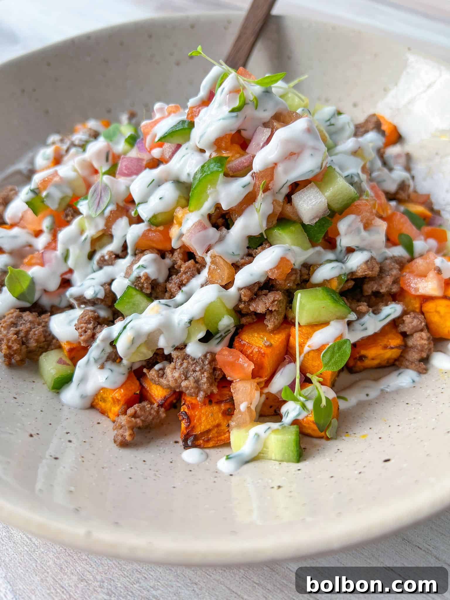 Sweet Potato Harvest Bowl 2 A vibrant bowl featuring seasoned ground beef nestled atop a bed of roasted sweet potatoes, generously topped with diced tomatoes, crisp cucumbers, and a creamy white sauce drizzle. Fresh microgreens garnish the dish, inviting a delightful culinary experience, all presented on a beige plate.