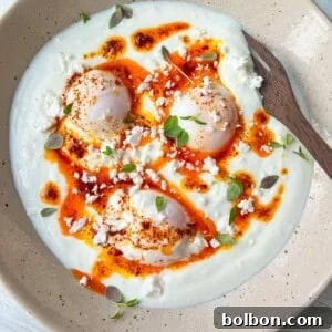 A dish featuring poached eggs on a bed of yogurt, drizzled with spicy red oil, garnished with crumbled feta cheese and fresh herbs. A wooden spoon rests on the beige plate.