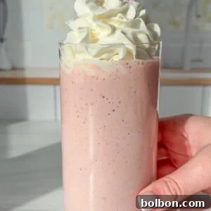 A hand holds a glass filled with a pink smoothie topped with a generous swirl of whipped cream. A red and white striped straw is placed in the drink. The smoothie is set against a light, blurred kitchen background.