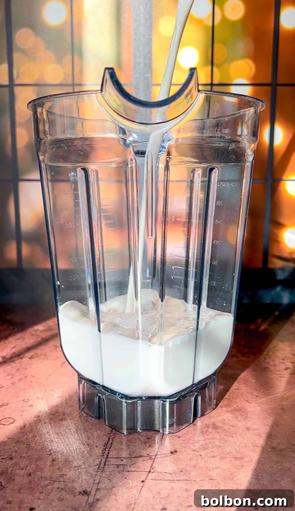 A blender pitcher being filled with milk, set on a wooden surface. The background is blurred with warm, golden bokeh lights, giving the scene a cozy and inviting atmosphere.