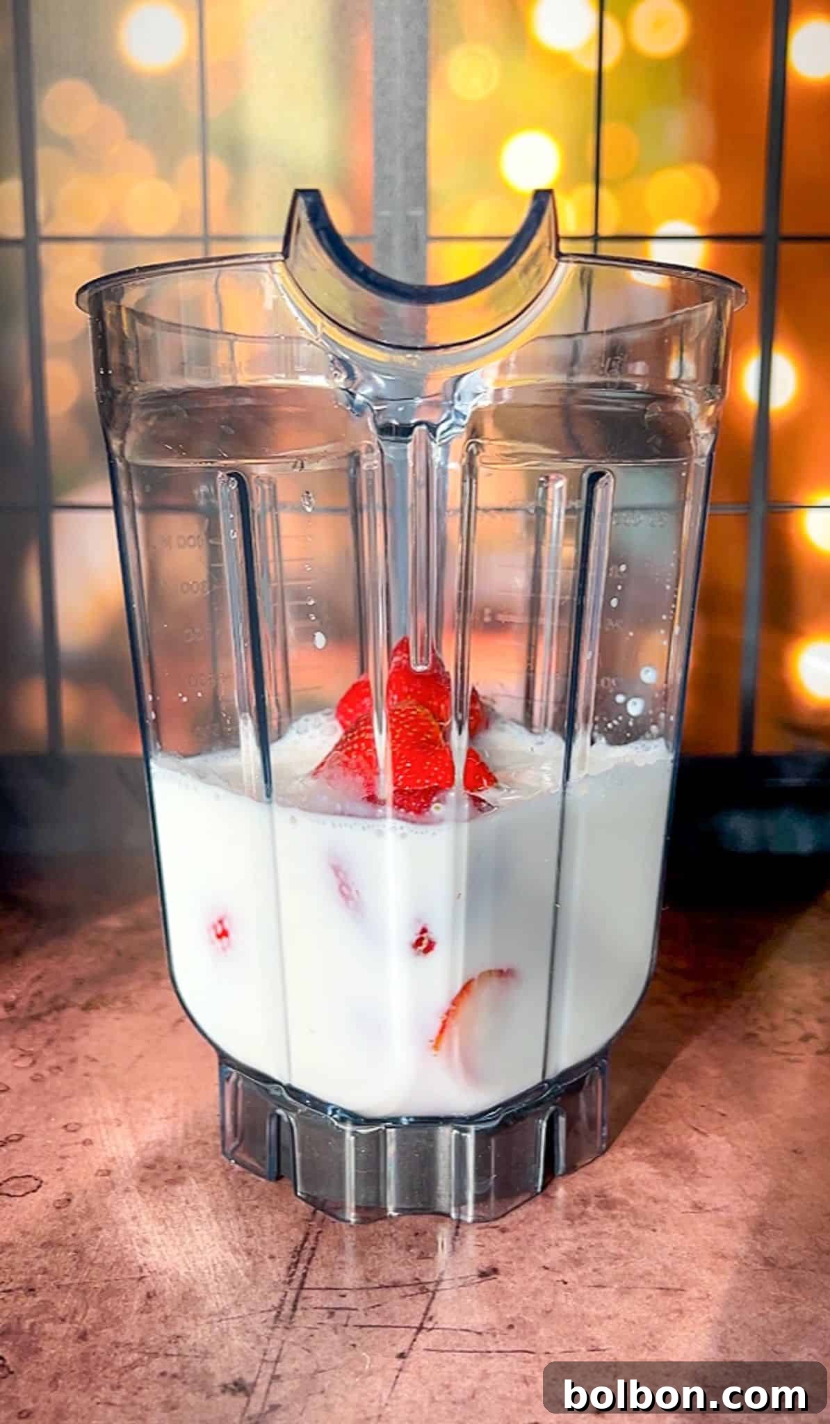 A blender jar filled with milk and sliced strawberries sits on a wooden surface. The background features warm, out-of-focus lights, creating a cozy atmosphere.
