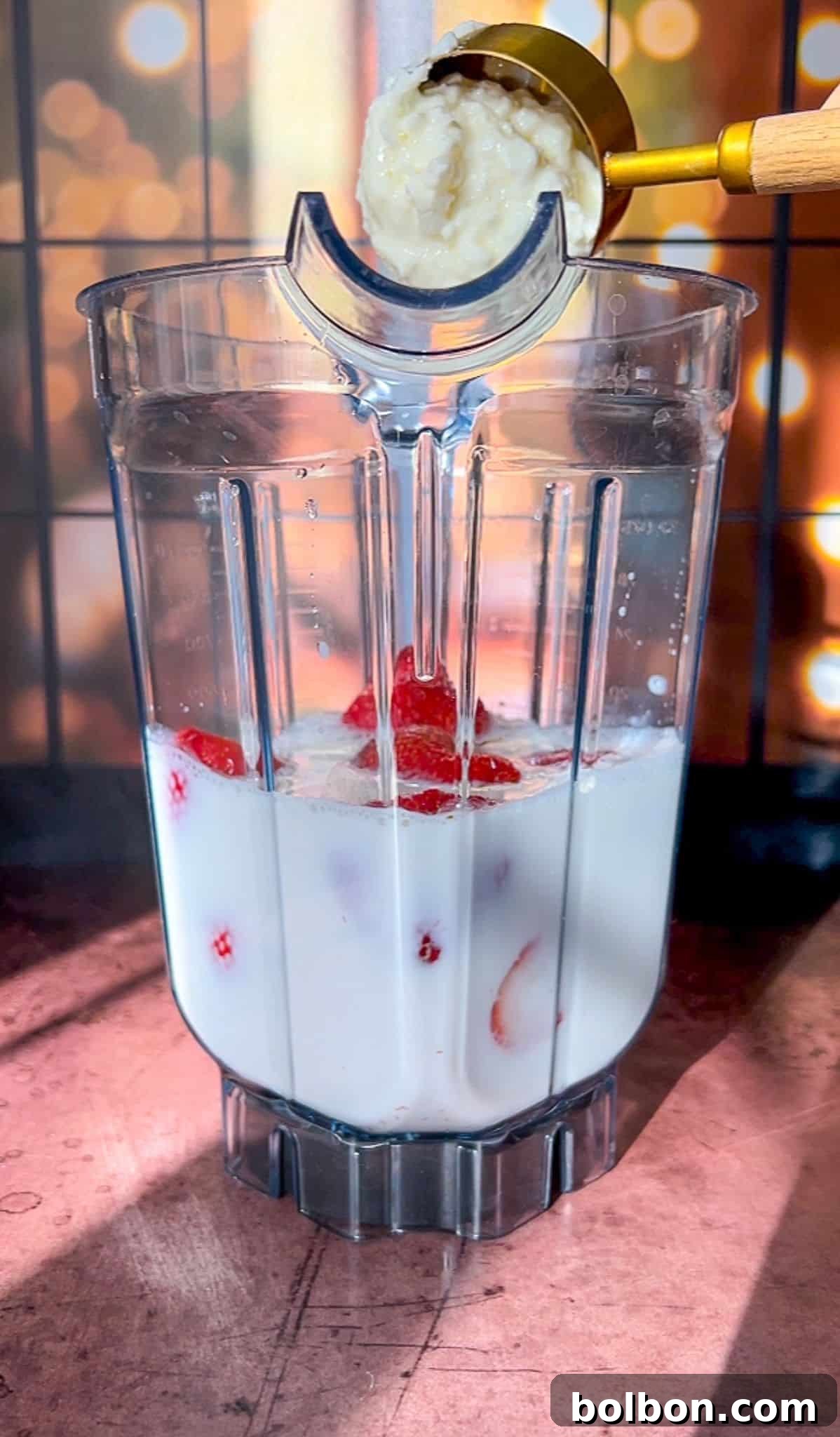 A blender jug filled with milk and strawberry slices sits on a countertop. A golden measuring cup pours a thick mixture, possibly banana puree, into the blender. The background is softly blurred with warm lights.