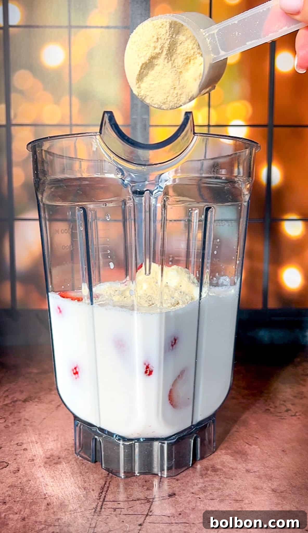 A blender pitcher filled with milk and strawberries on a wooden surface. A hand is adding a scoop of brown powder into the blender. The background features warm, blurred lights creating a cozy ambiance.