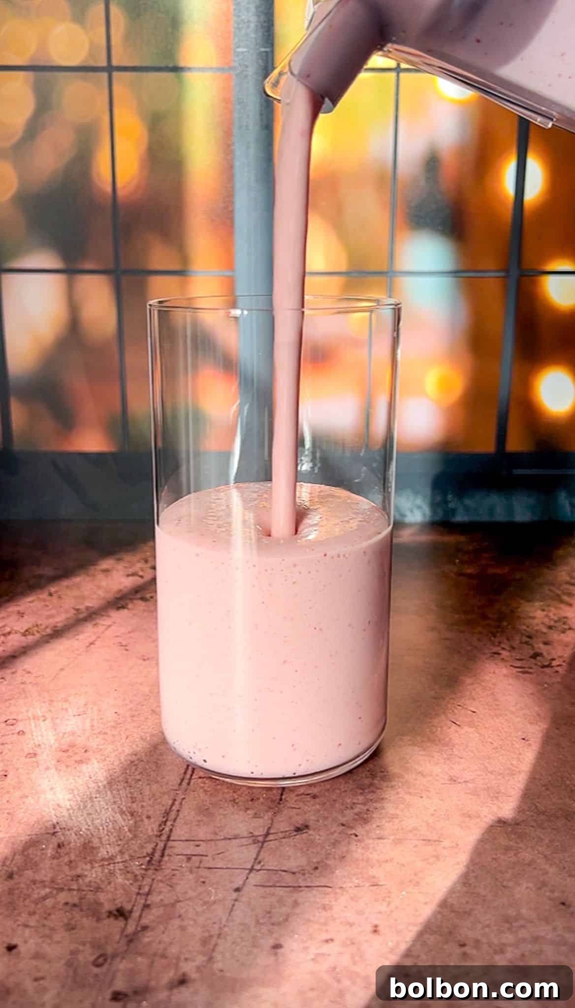 A tall glass on a wooden table is being filled with a strawberry protein shake being poured from a blender. The background has warm bokeh lights and a grid-patterned window, creating a cozy atmosphere.