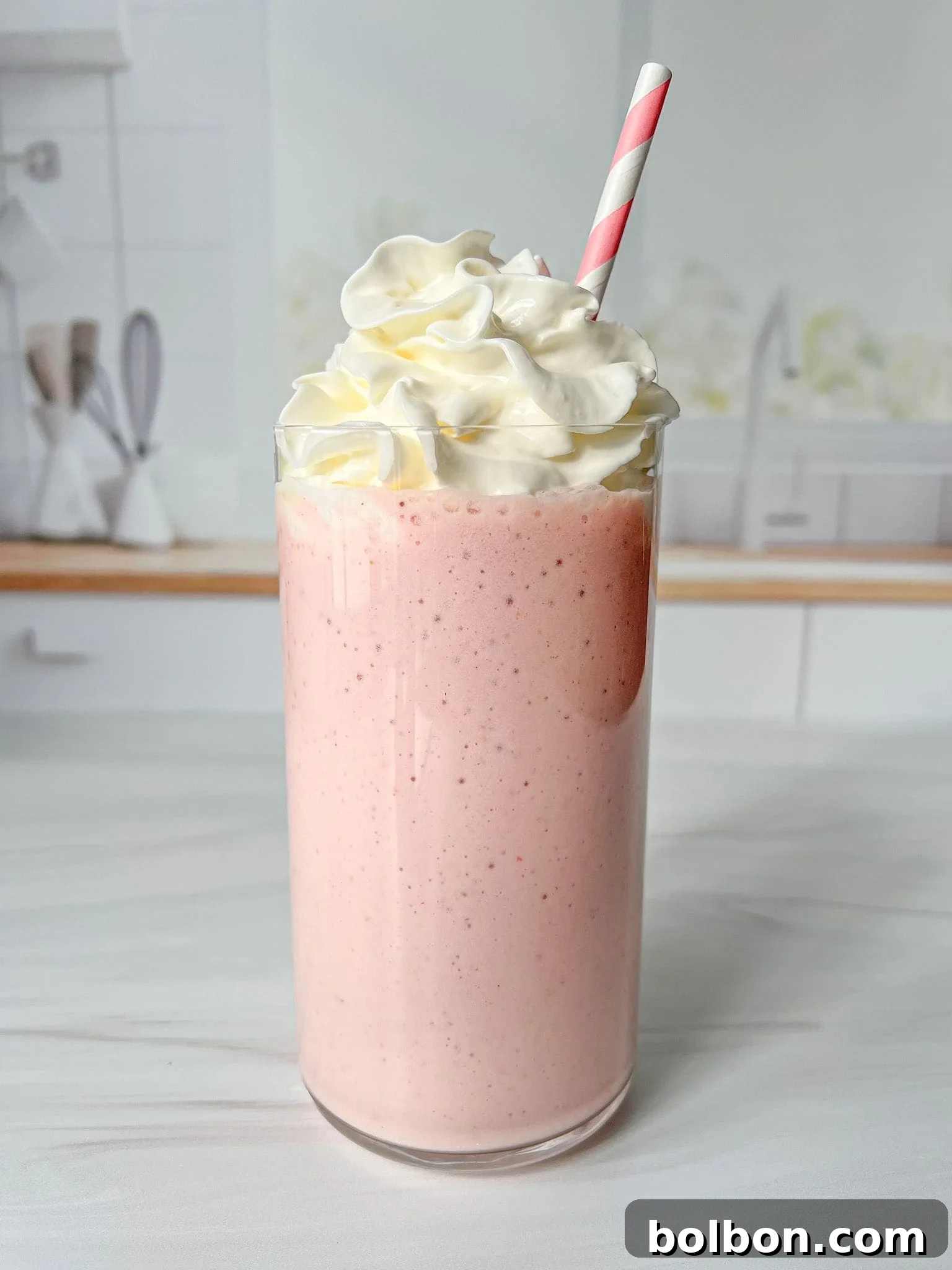A tall glass filled with a strawberry protein shake topped with swirls of whipped cream, and a pink and white striped straw. The background shows a blurred kitchen setting.