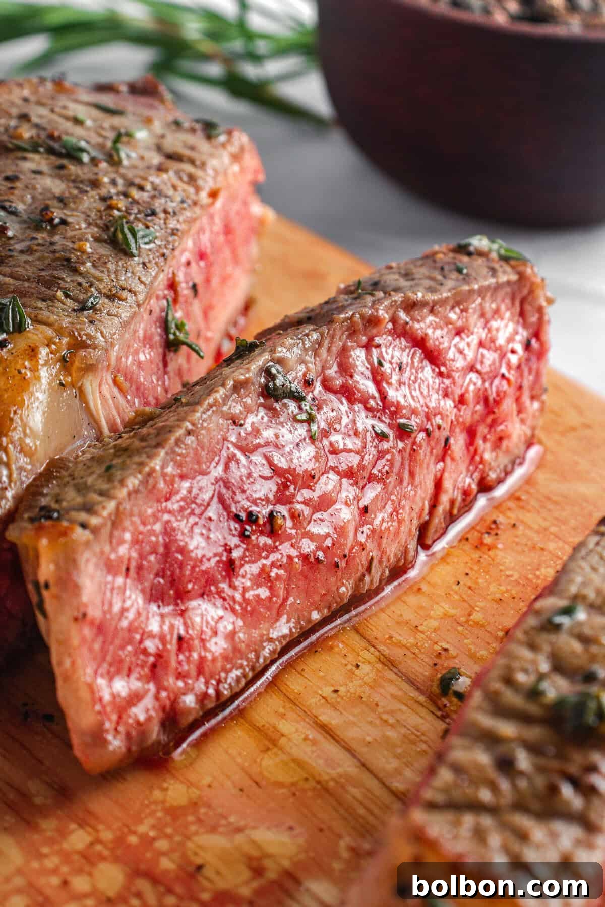 Close-up of juicy, medium-rare steak slices on a wooden cutting board, garnished with herbs and pepper. A blurred bowl with herbs is in the background.