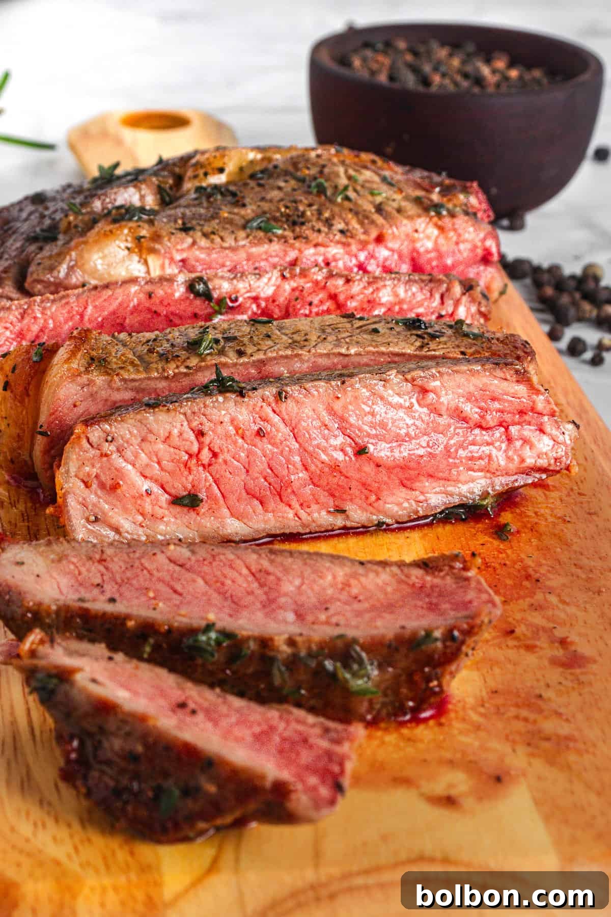 Slices of medium-rare steak on a wooden cutting board, garnished with herbs. In the background, a black bowl filled with peppercorns and a wooden spoon are visible. The steak is juicy with a seared crust and pink interior.