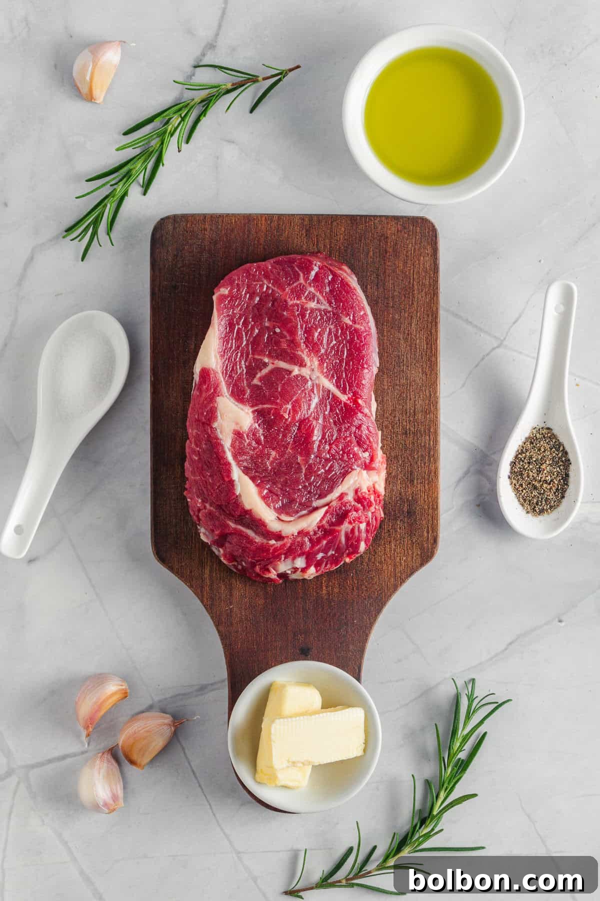 A raw steak on a wooden cutting board surrounded by small bowls of olive oil, butter, garlic cloves, rosemary sprigs, and spoons of salt and pepper, all placed on a light marble surface.