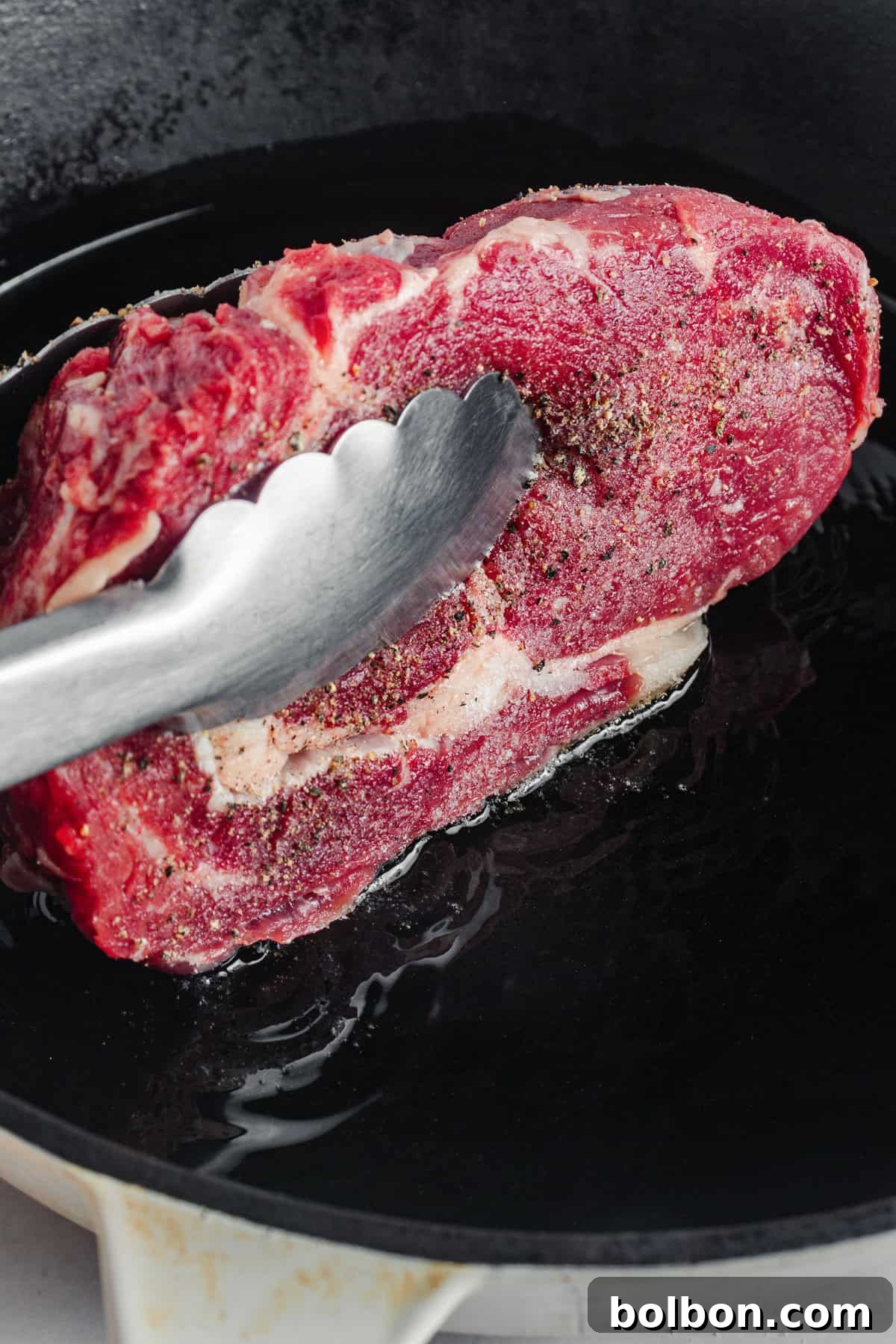A raw, marbled steak seasoned with pepper is being placed into a hot cast iron skillet using metal tongs. The steak appears to be in the initial stages of cooking.