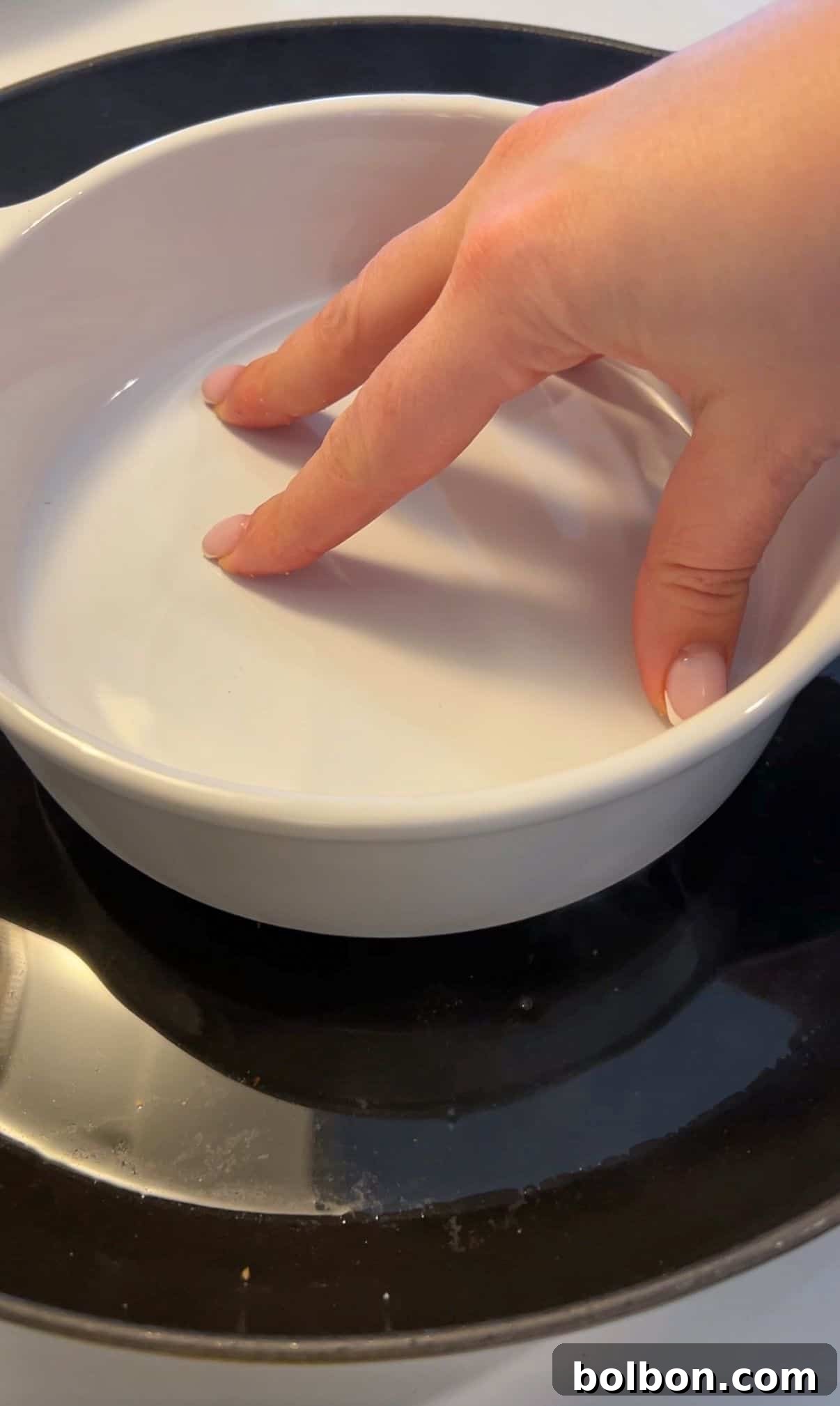 A hand with light pink nails is gently touching the inside of a white ceramic dish, which is placed on a black stovetop.