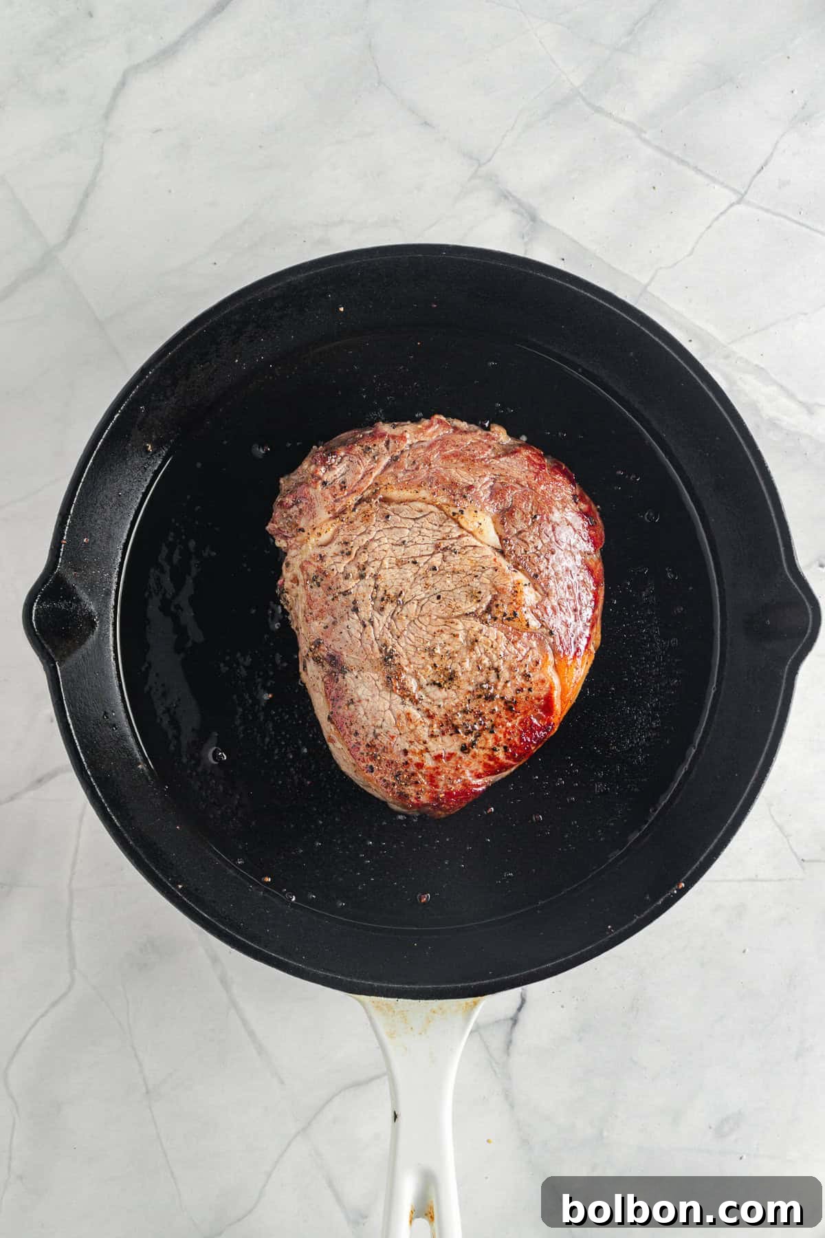 A piece of seared meat cooking in a black cast iron skillet on a gray marble surface. The meat has a browned exterior, and the pan shows traces of oil.