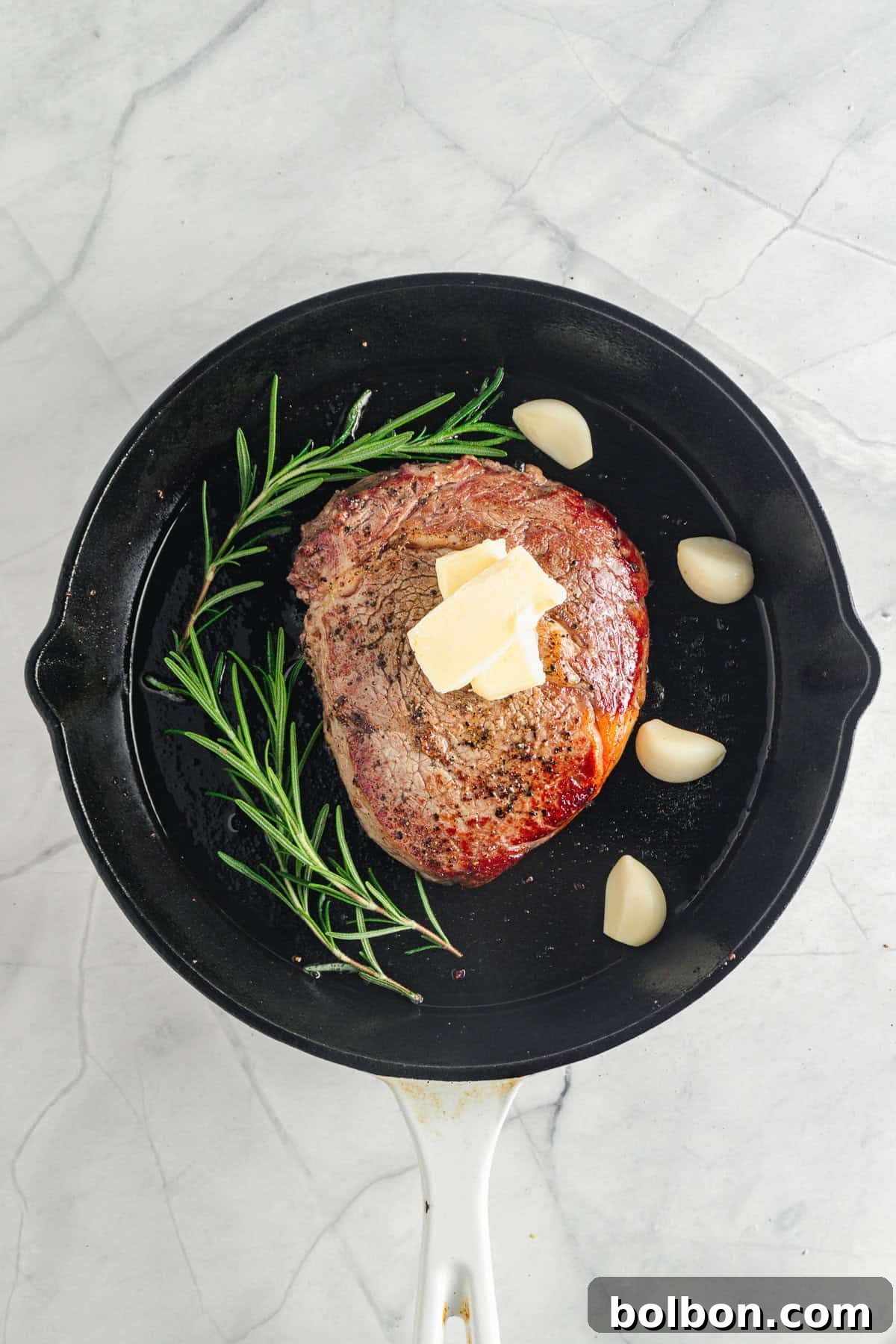 The seasoned meat topped with butter is in a black frying pan. Sprigs of rosemary and garlic cloves surround it. The pan sits on a light marble surface.