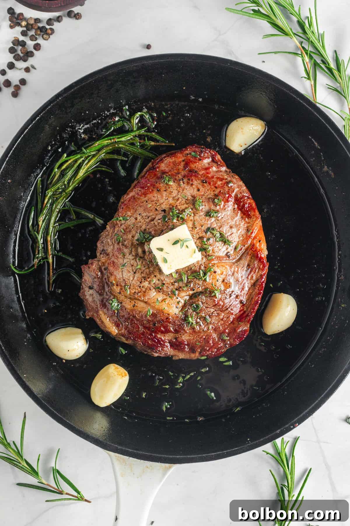 The grilled meat topped with a pat of butter and sprinkled with herbs sits in a skillet. Surrounding the meat are cloves of garlic and sprigs of rosemary.