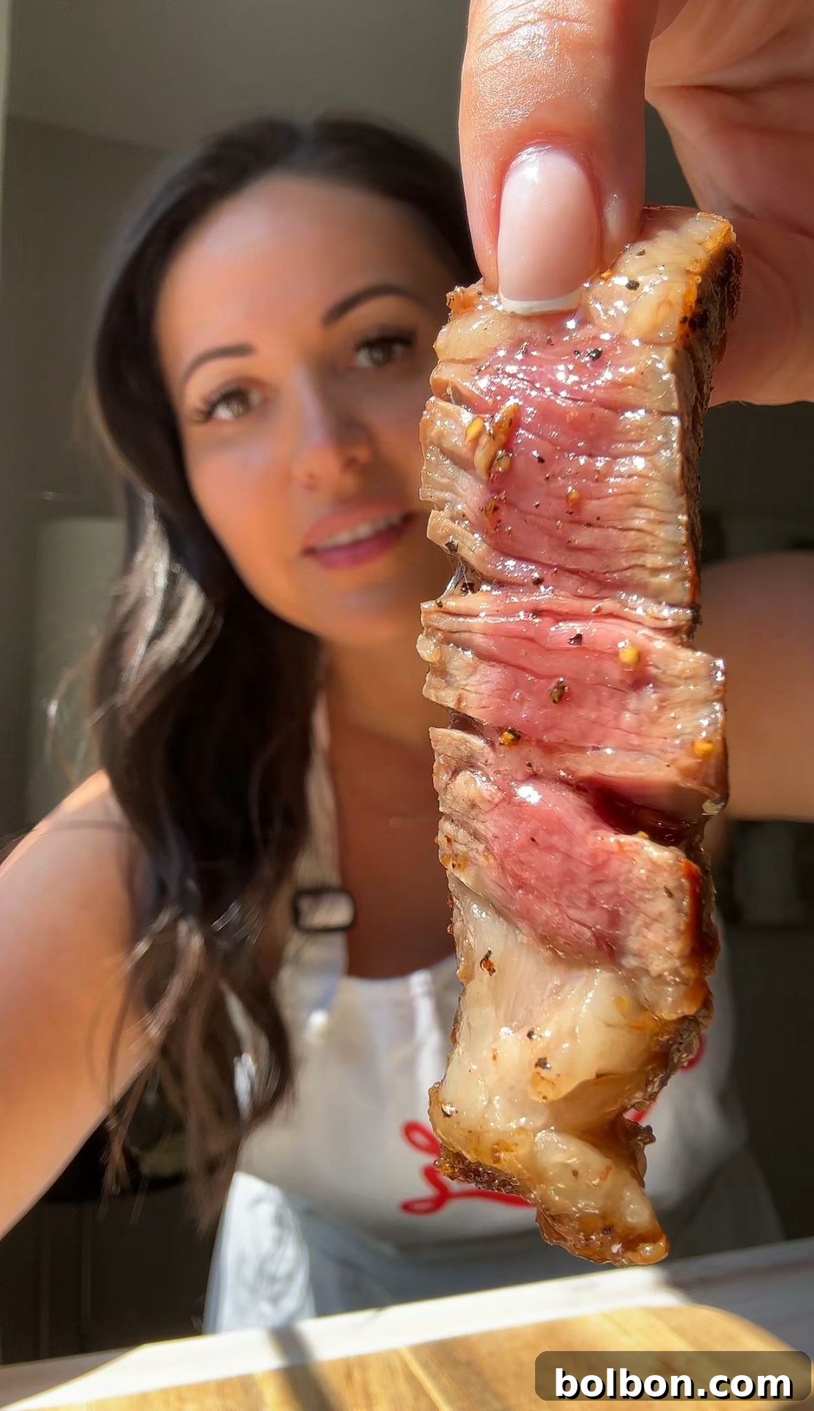 A woman holds up a juicy, grilled steak slice, showing the medium-rare interior. She is smiling and wearing a white apron. The background is softly blurred, highlighting the focus on the steak.