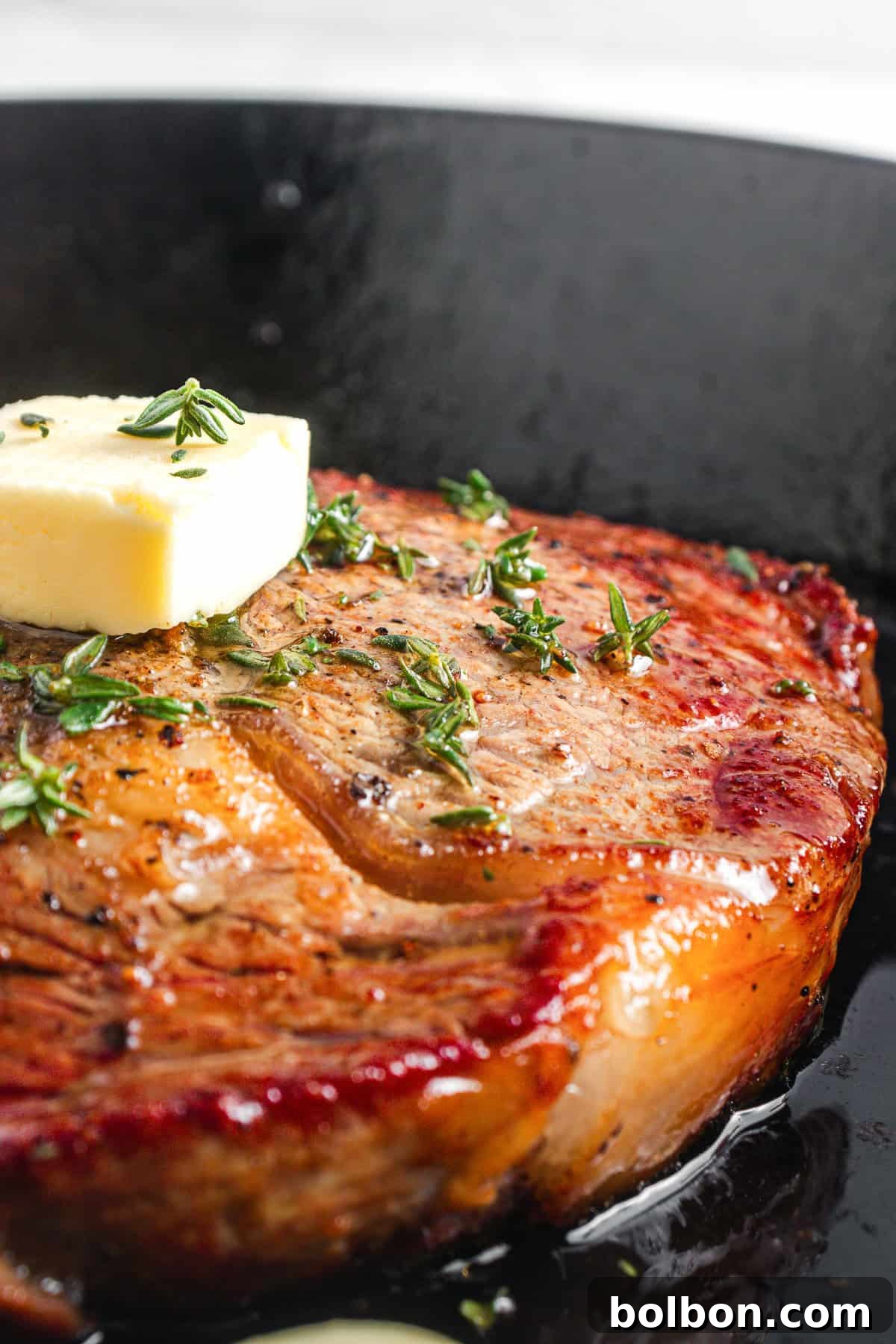 A close-up of a seared steak in a black skillet, topped with a melting pat of butter and sprinkled with fresh thyme. The steak is browned and glistening, with visible grill marks and a juicy appearance.