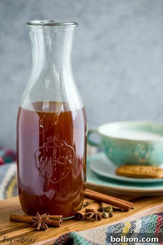 A large glass bottle filled with rich, homemade chai concentrate, surrounded by whole spices like cinnamon sticks and cardamom pods