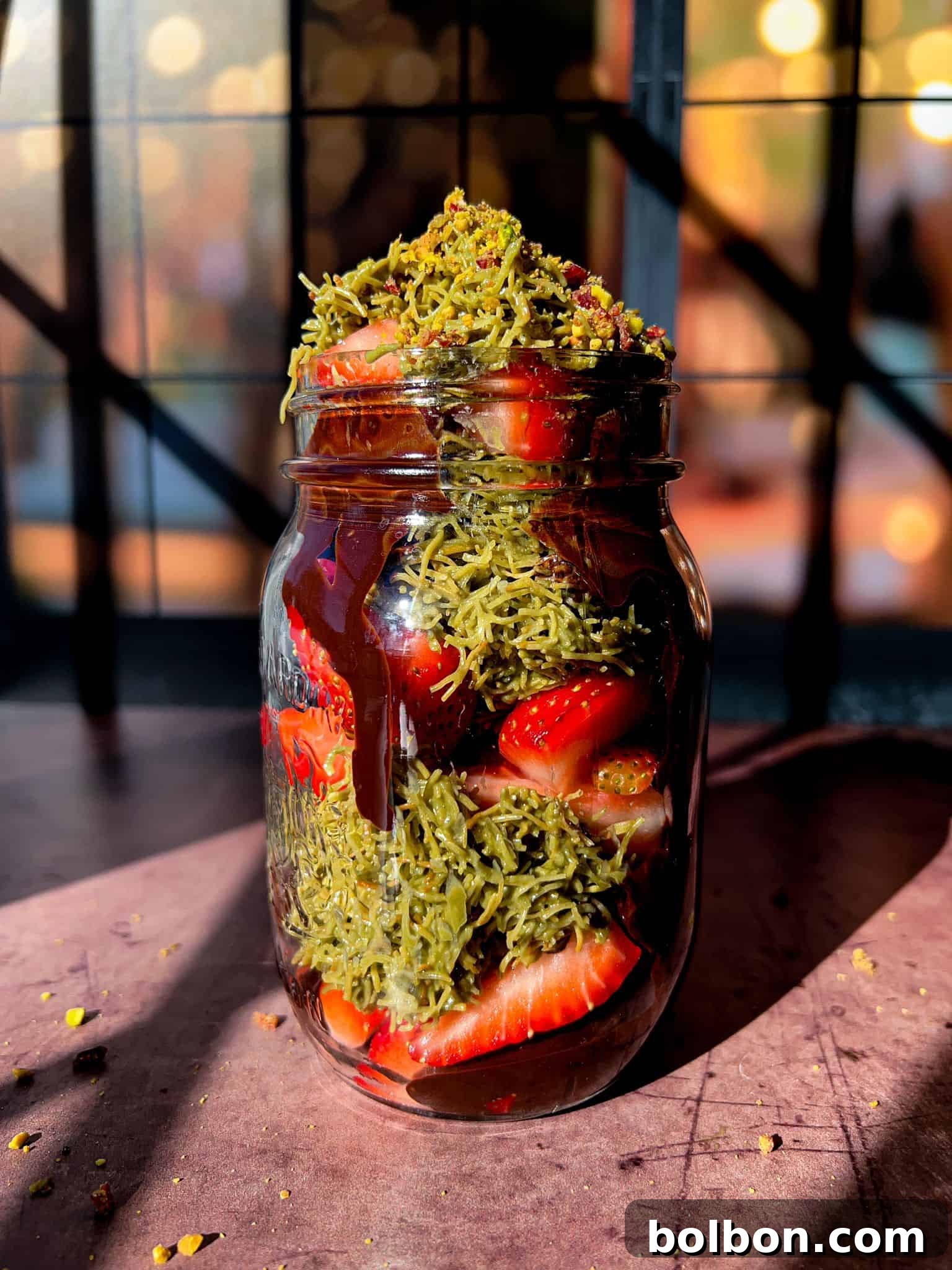 The Dubai Strawberry cup, a layered dessert presented in a clear jar, features alternating layers of fresh sliced strawberries, smooth melted chocolate, and a vibrant green shredded topping, likely pistachios. The dessert sits on a light surface with a soft, blurred background, highlighting its appetizing appearance.