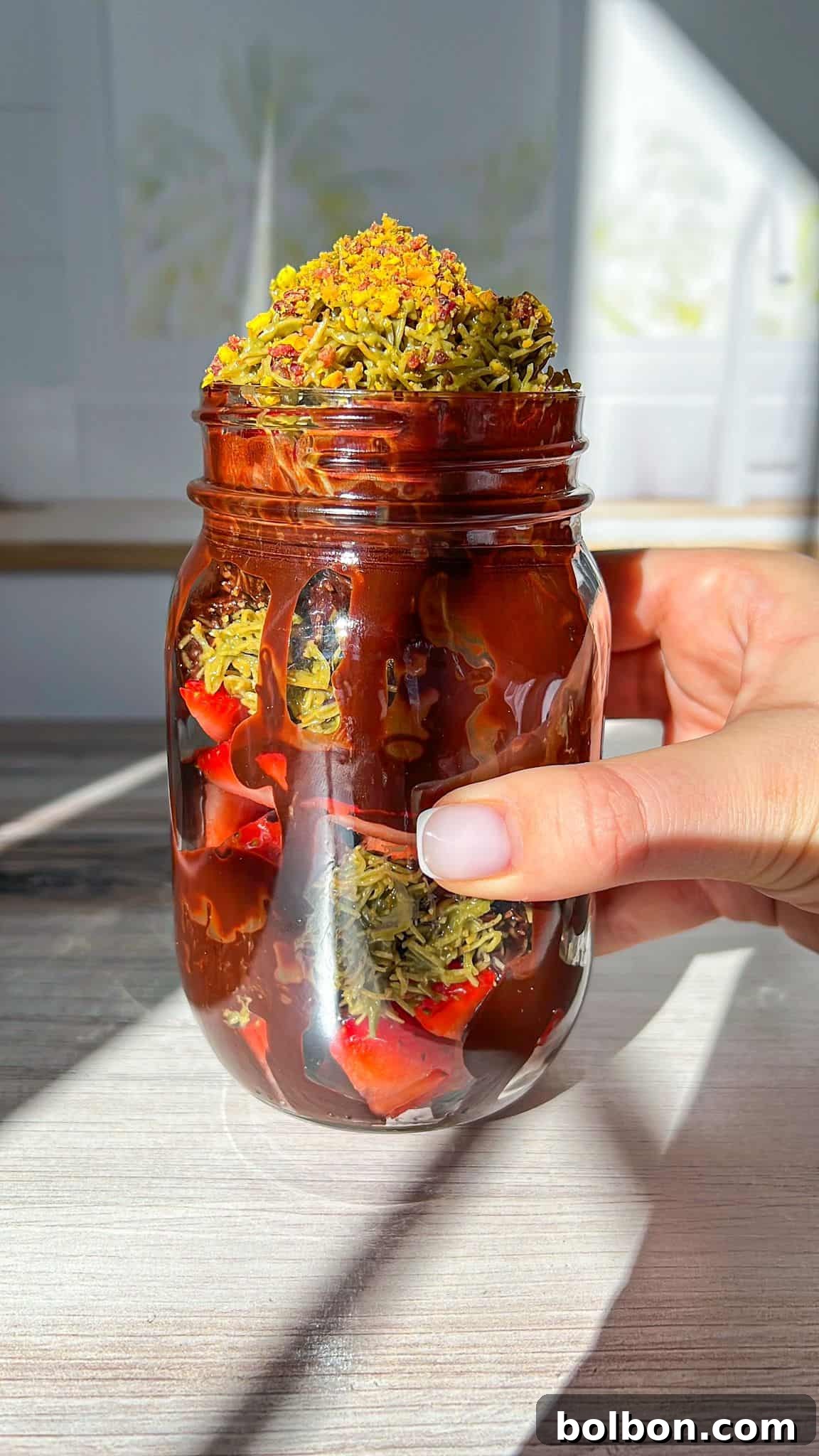 A hand gently holds a clear glass jar filled with the beautifully layered Dubai Strawberry Cup. The dessert features visible layers of vibrant red strawberries, rich chocolate sauce, and crumbled green pistachio topping. The jar is set against a textured wooden surface, illuminated by natural sunlight that creates warm shadows.