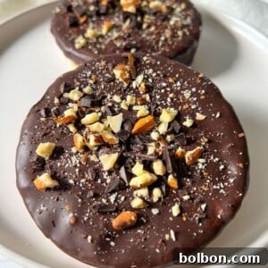 Two round Chocolate Covered Rice Cakes topped with chopped nuts and chocolate pieces sit on a white plate with a light background.