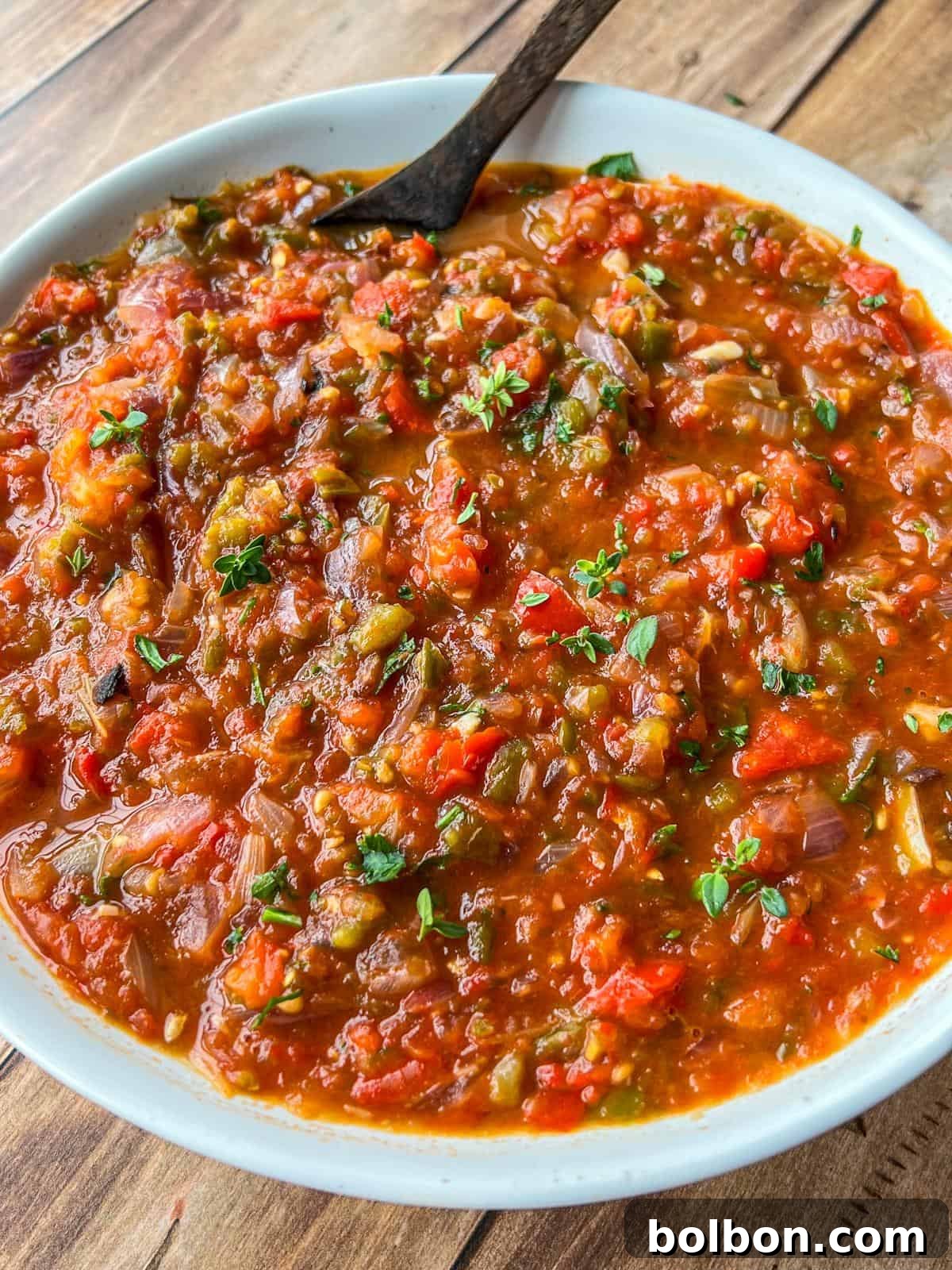 A close-up of a bowl filled with chunky, homemade roasted vegetable salsa, featuring visible pieces of tomato, pepper, onion, and herbs, with a spoon resting inside the bowl on a wooden table.