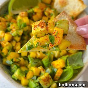 A close-up of a hand holding a chip topped with mango salsa, featuring diced mango, avocado, herbs, and spices. A bowl of the fresh Mango Salsa Recipe and a lime wedge are visible in the background.
