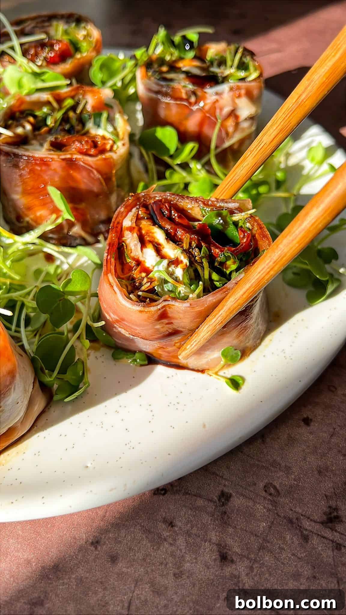 A close-up of bacon-wrapped rolls filled with spinach and sun-dried tomatoes, garnished with microgreens on a white plate. Chopsticks hold one roll. The setting is bright with natural light.