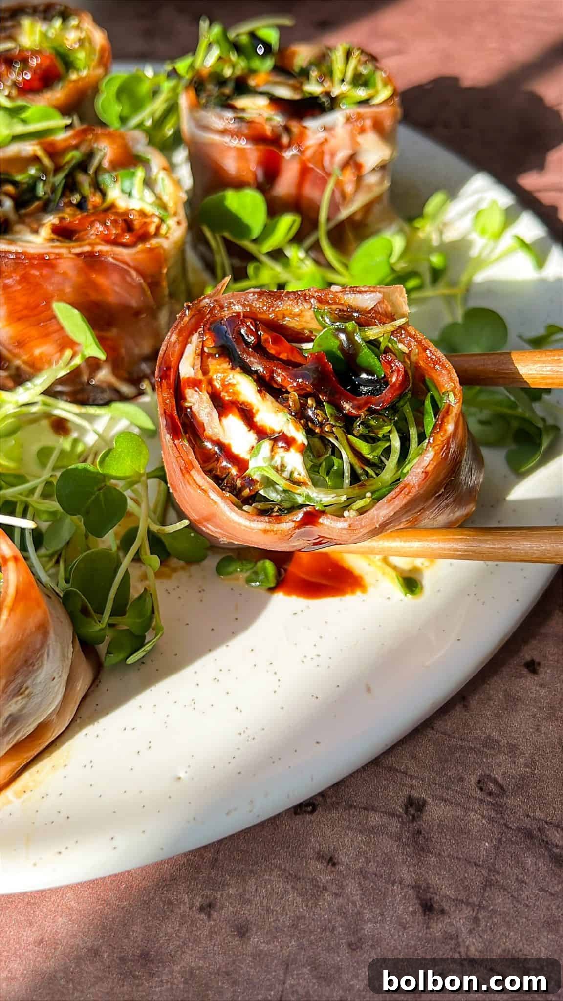 Close-up of Italian sushi recipe wrapped in prosciutto, filled with greens, and drizzled with a dark sauce on a white plate. They're accompanied by small leafy greens, with chopsticks on the side. The setting is warmly lit.