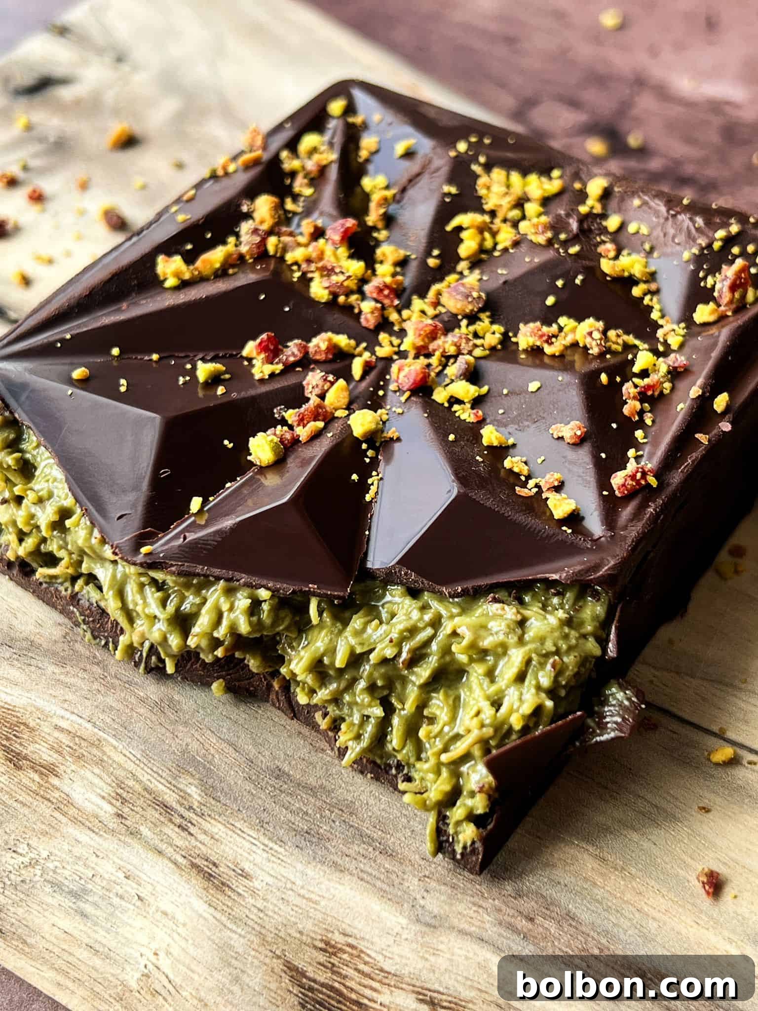 Desert Gold Chocolate Bar 2 A chocolate dessert with a geometric pattern on top, filled with green matcha cream. Crumbled yellow and red toppings are scattered on top, and the dessert sits on a wooden surface.