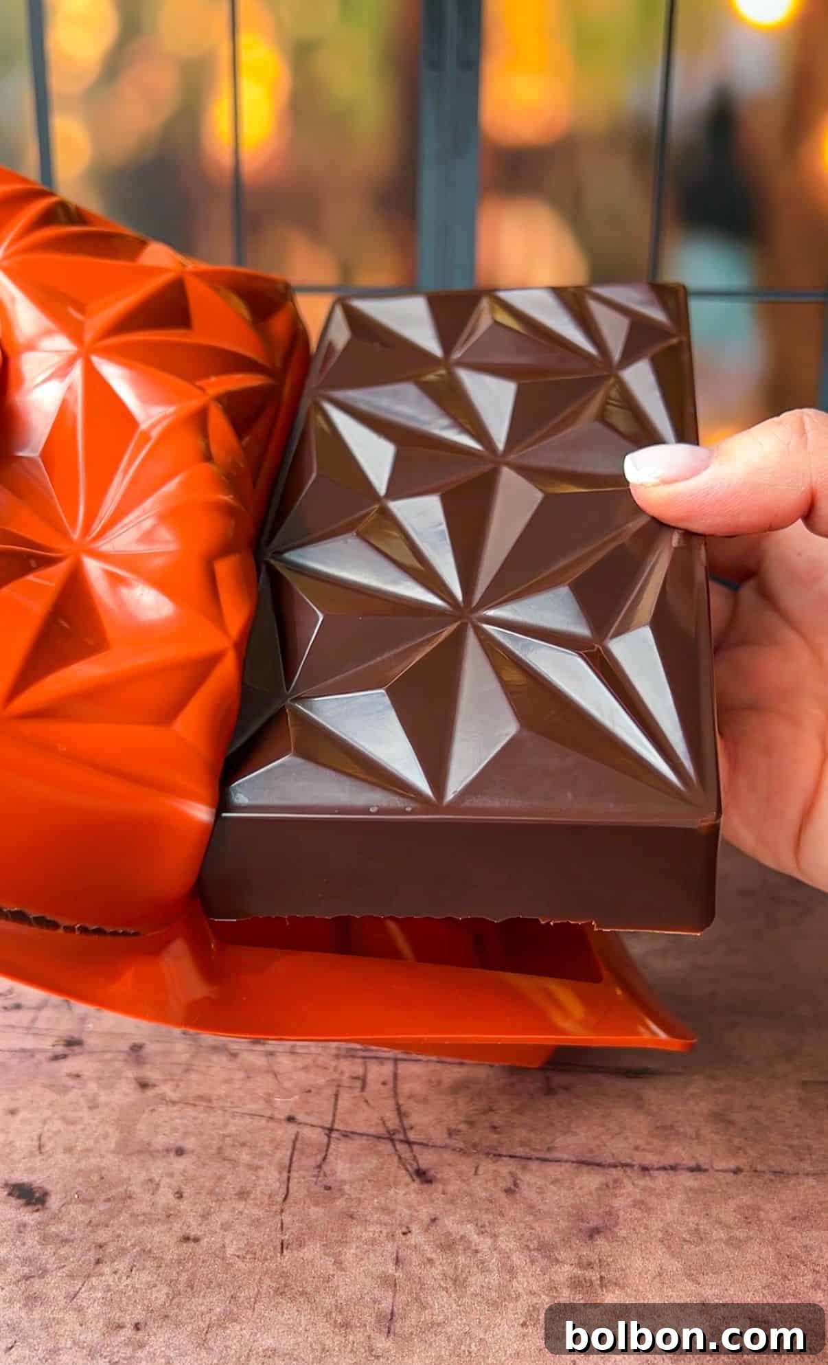 Desert Gold Chocolate Bar 11 A geometric chocolate bar is being lifted by a hand, showcasing a pattern of triangles. The dark brown chocolate contrasts with an orange mold in the background. The setting appears warm and rustic.