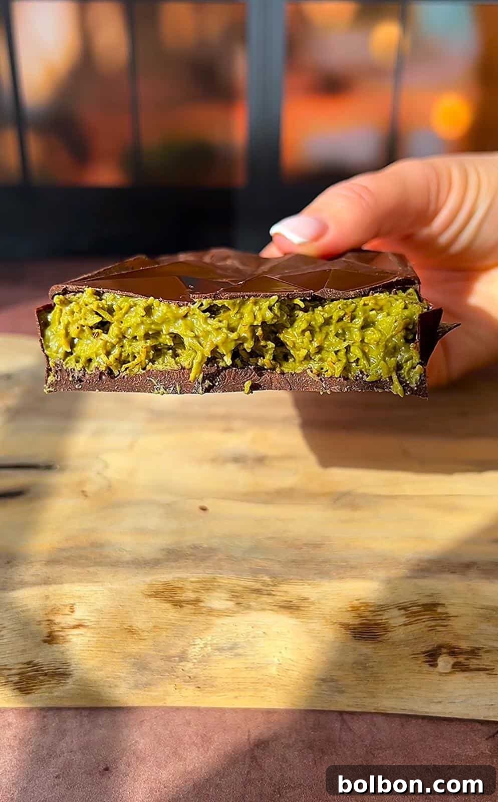 Desert Gold Chocolate Bar 12 A close-up of a hand holding a cross-section of a chocolate bar with a green filling. The filling has a textured, fibrous appearance, and the bar is resting on a wooden surface with a blurred background.