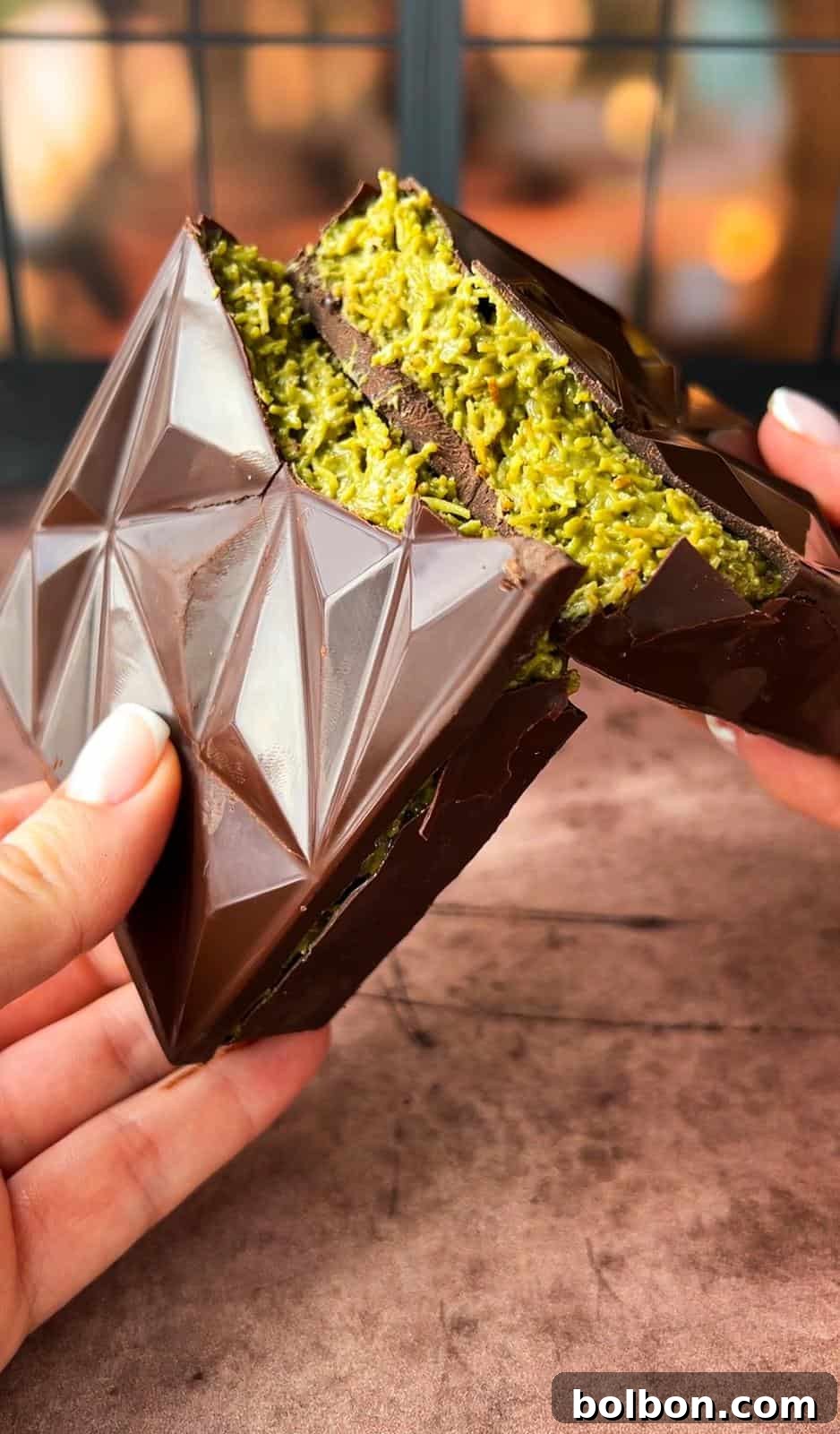 Desert Gold Chocolate Bar 13 Two hands hold a chocolate dessert filled with a bright green, shredded filling. The chocolate has a geometric pattern, and the image is set against a blurred background with dark framing.