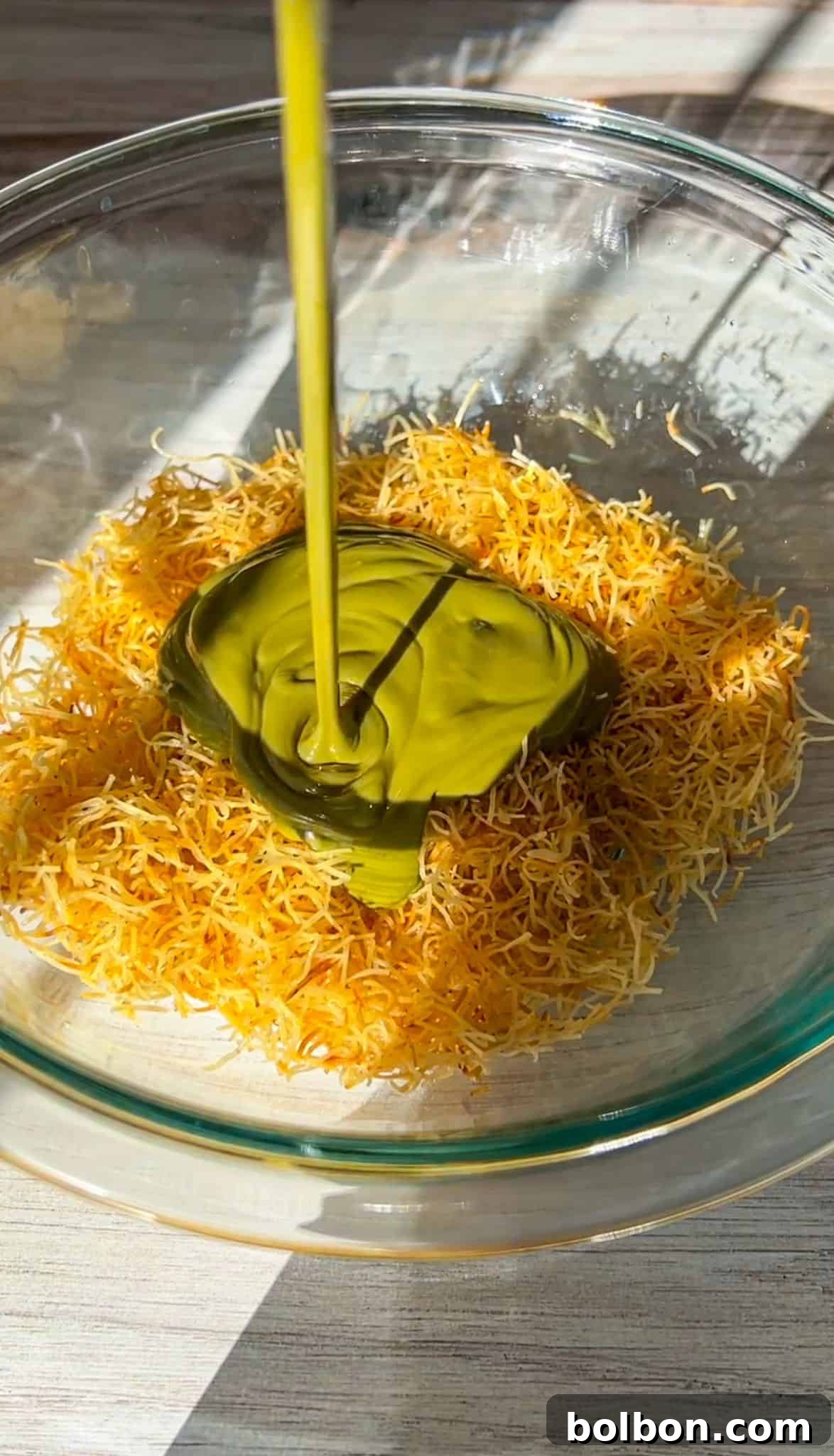 Desert Gold Chocolate Bar 6 Shredded pastry dough is placed in a glass bowl with a green paste on top. A stream of golden liquid is being poured over the mixture. Bright sunlight is casting shadows on the surface.