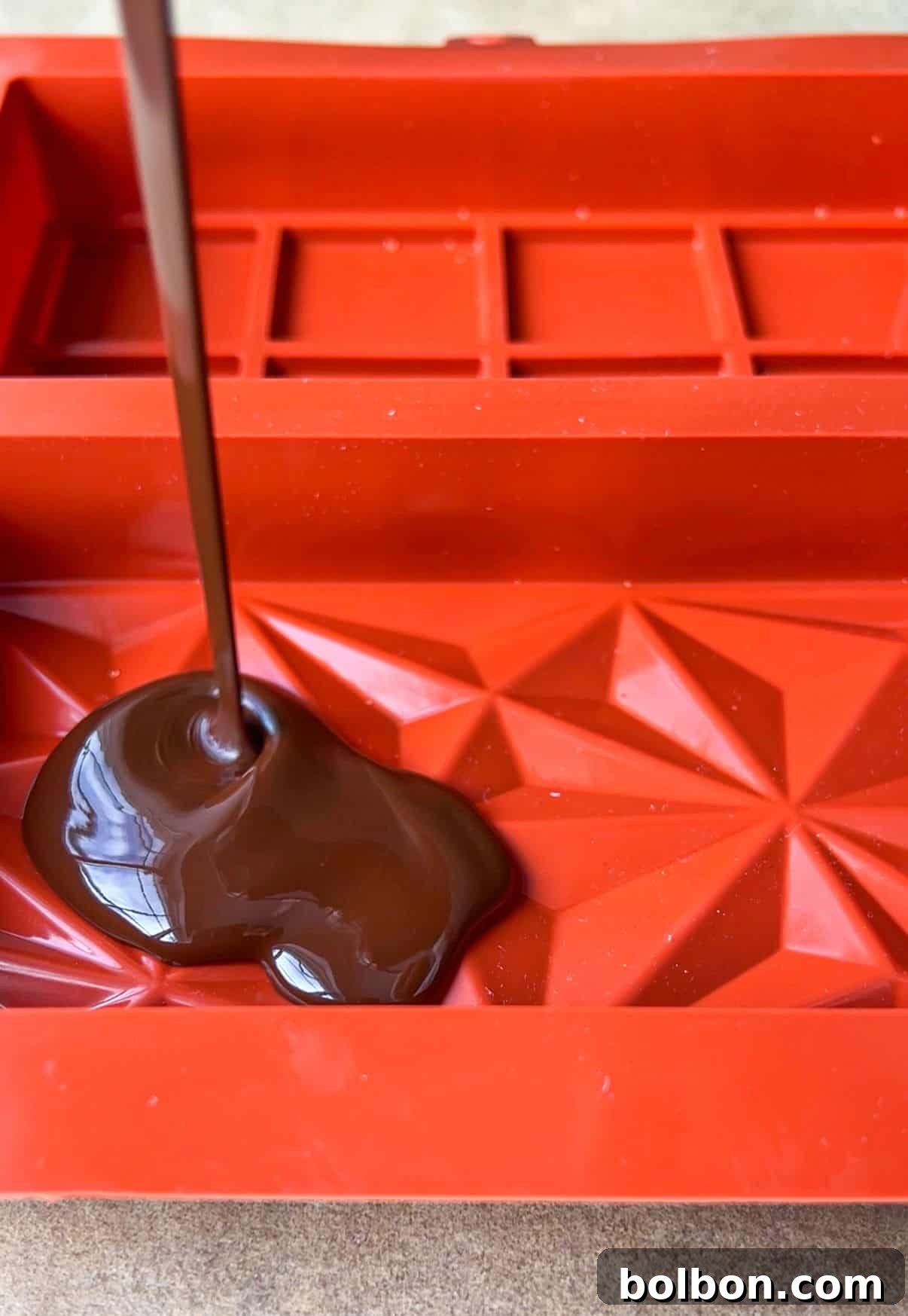 Desert Gold Chocolate Bar 8 Melted chocolate being poured into a red silicone mold with geometric patterns and rectangular sections.