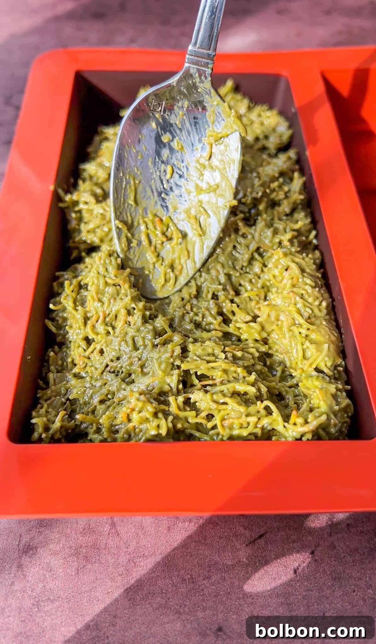 Desert Gold Chocolate Bar 9 A close-up of a rectangular red silicone mold filled with a green mixture. A metal spoon is spreading the mixture evenly across the mold's surface. The background is a textured surface in a pinkish hue.