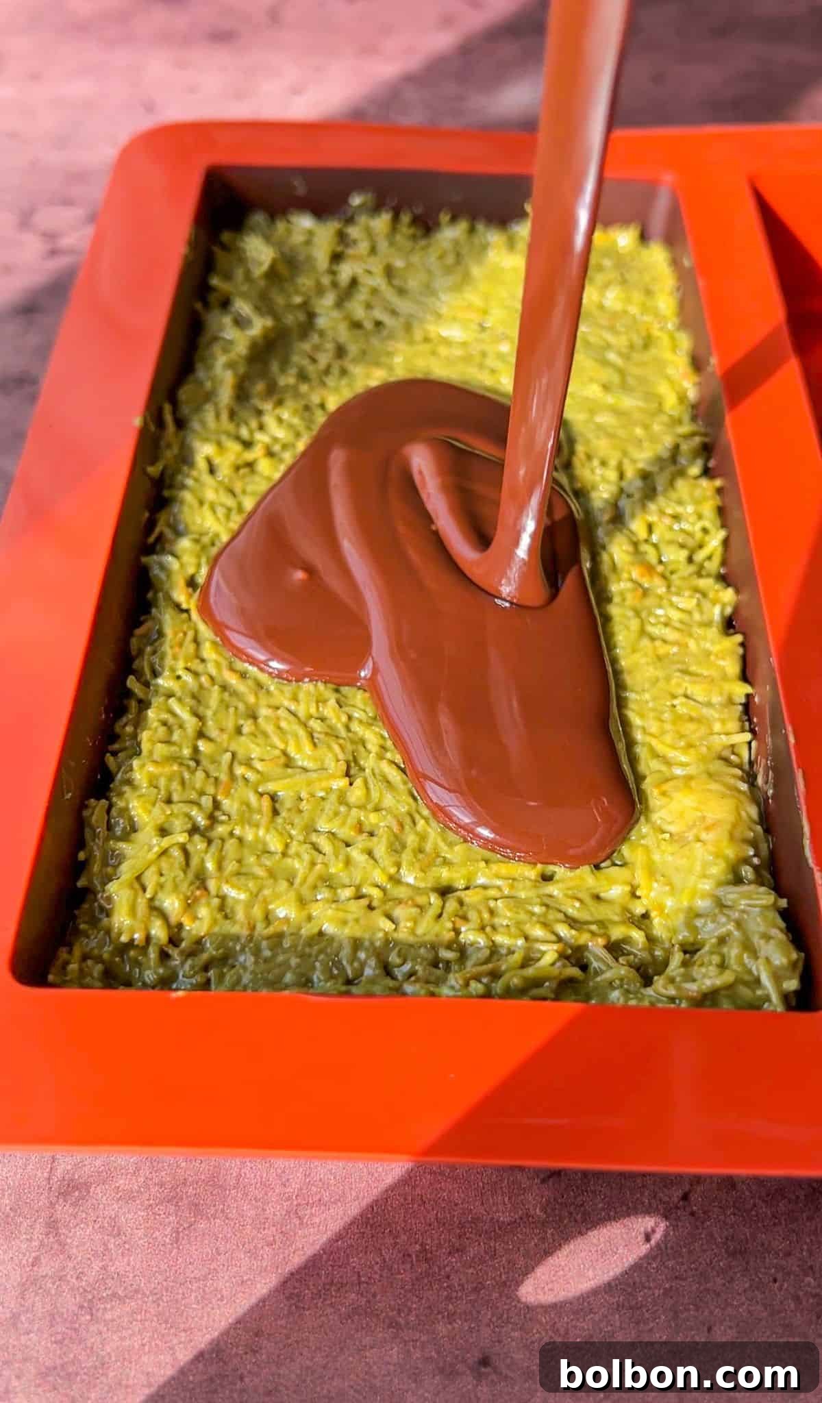 Desert Gold Chocolate Bar 10 Chocolate being poured over a block of green dessert in a red rectangular mold. The dessert appears to have a textured, shredded surface.