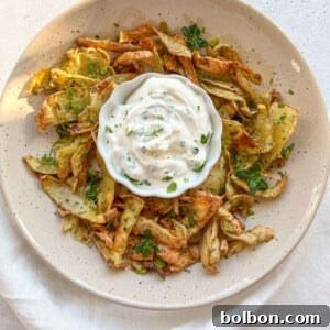 A beige plate with crispy vegetable chips garnished with herbs surrounds a bowl of creamy white dip, set on a white surface with a white napkin underneath.